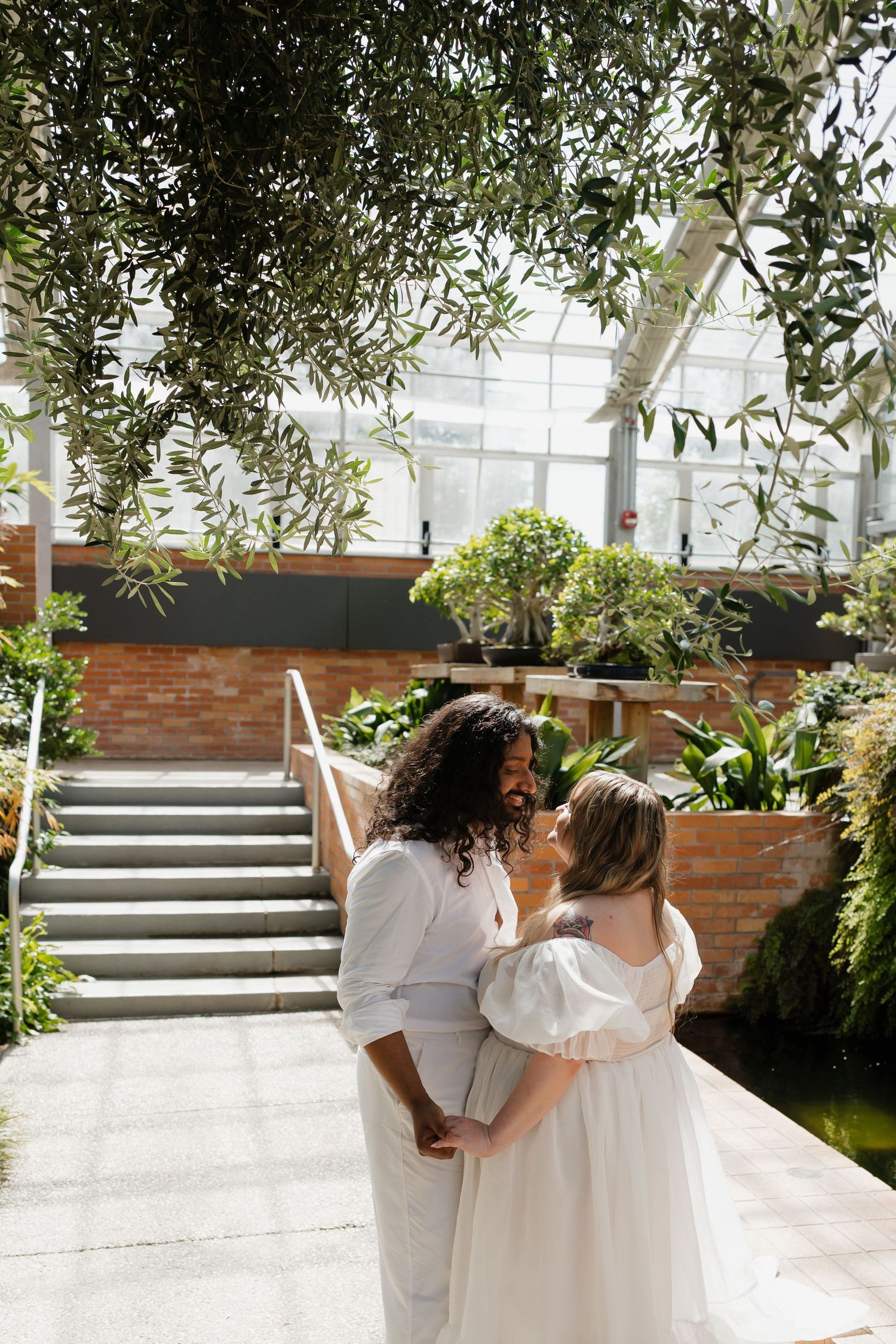 Interracial couple embracing and smiling at each other in a sunlit botanical garden greenhouse.