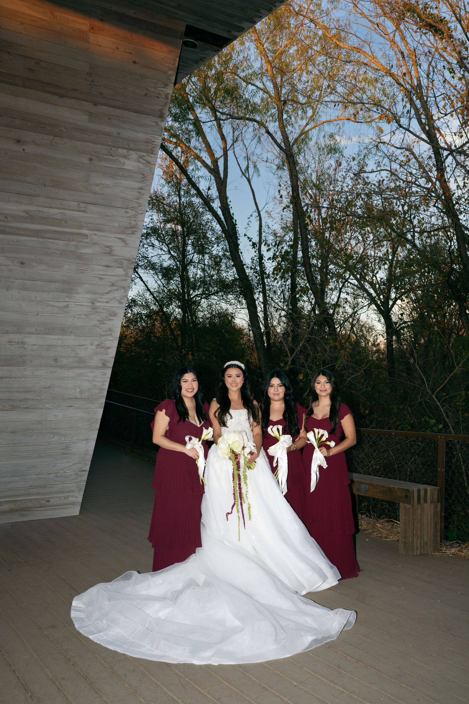 Bride in white gown with three bridesmaids in burgundy dresses holding bouquets at outdoor wedding venue.