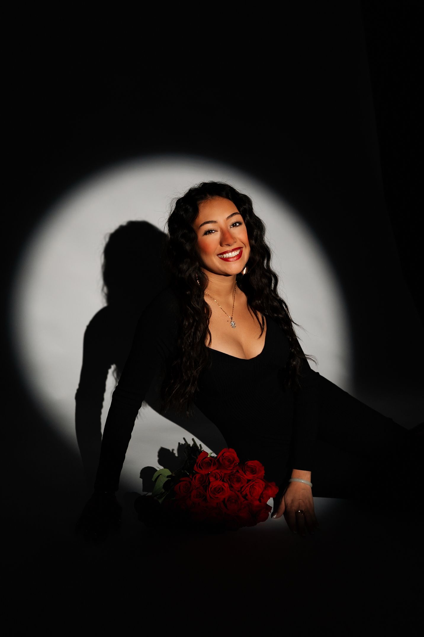 Smiling young woman with curly hair holding red roses in a dramatic black and white spotlight.