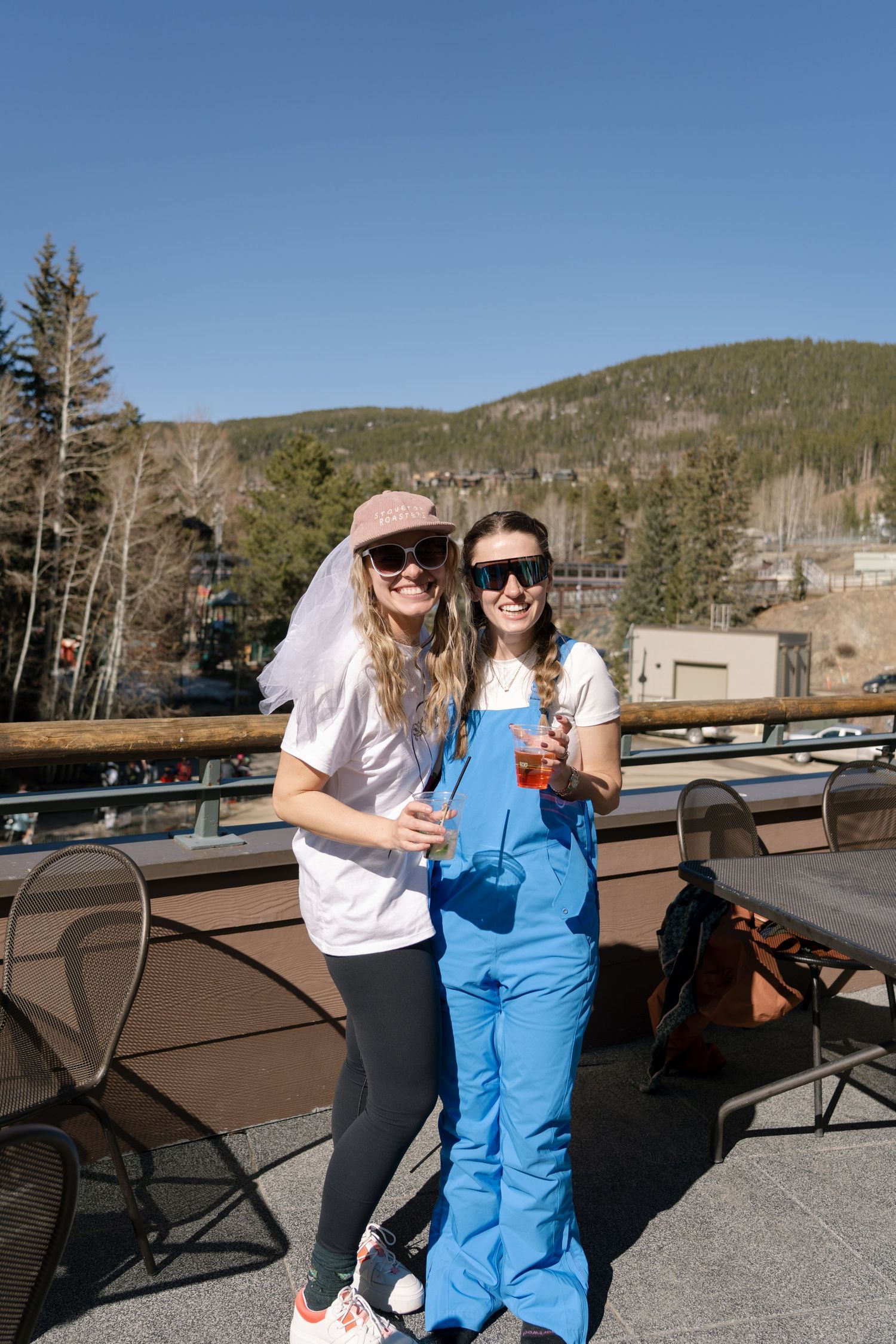 Two women in sunglasses holding drinks on a mountain resort deck with tree-covered hills behind.