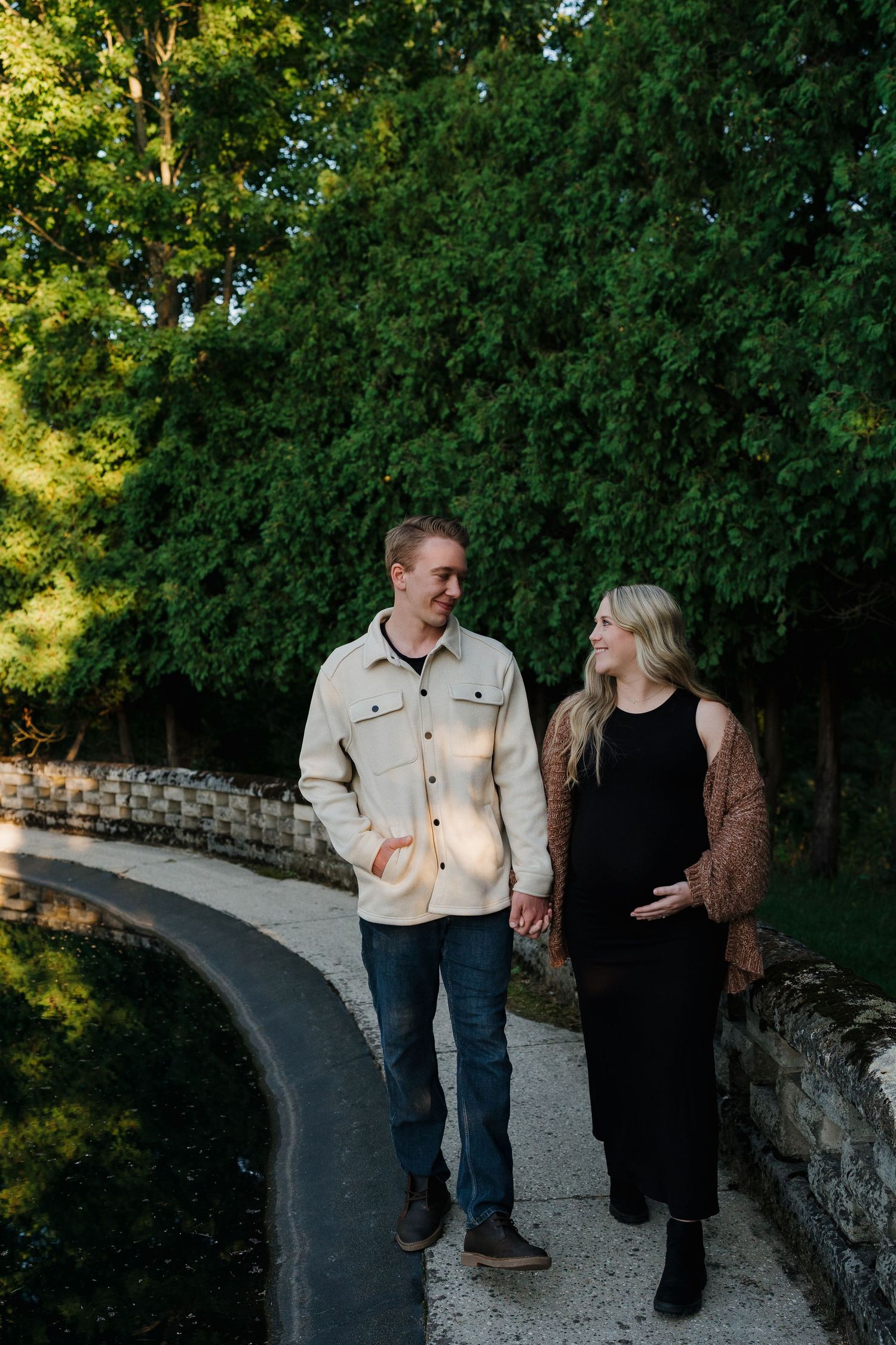 Couple holding hands walking along a stone bridge path surrounded by lush green trees during golden hour.