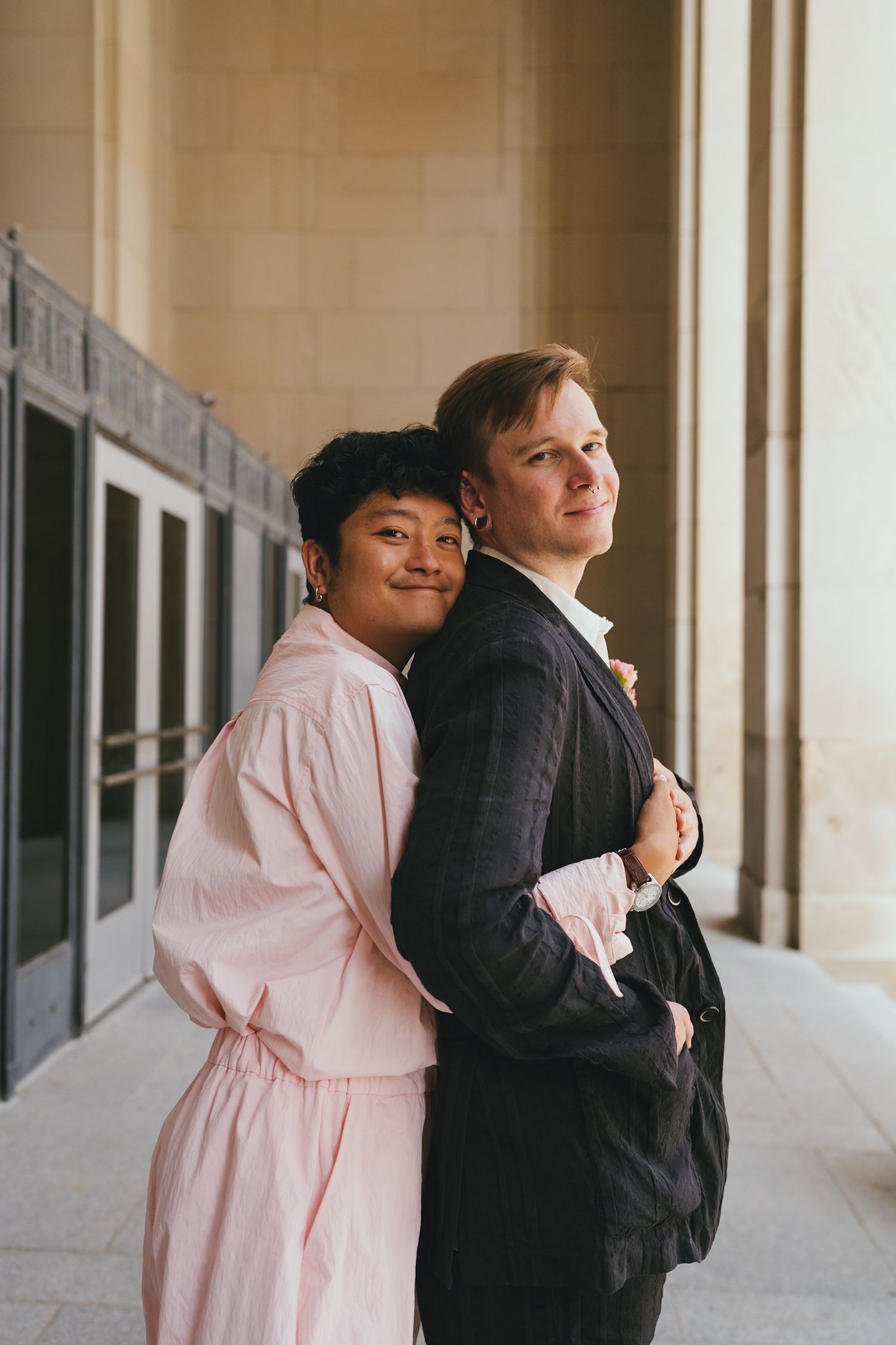 Couple standing back-to-back in corridor, woman in pink robe embracing man in dark suit, both smiling confidently.