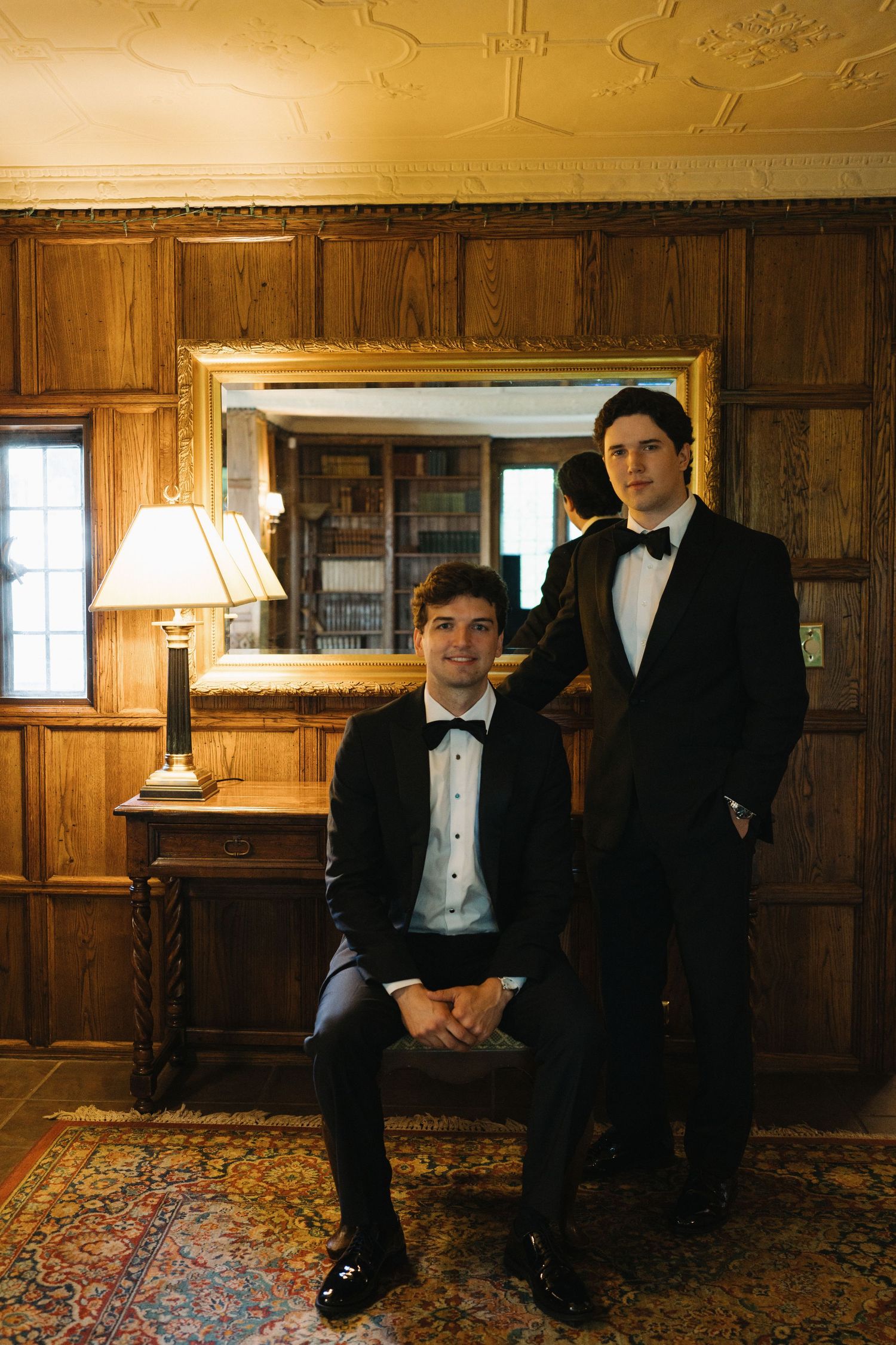 Two groomsmen in black tuxedos pose in an elegant wood-paneled room with vintage decor.