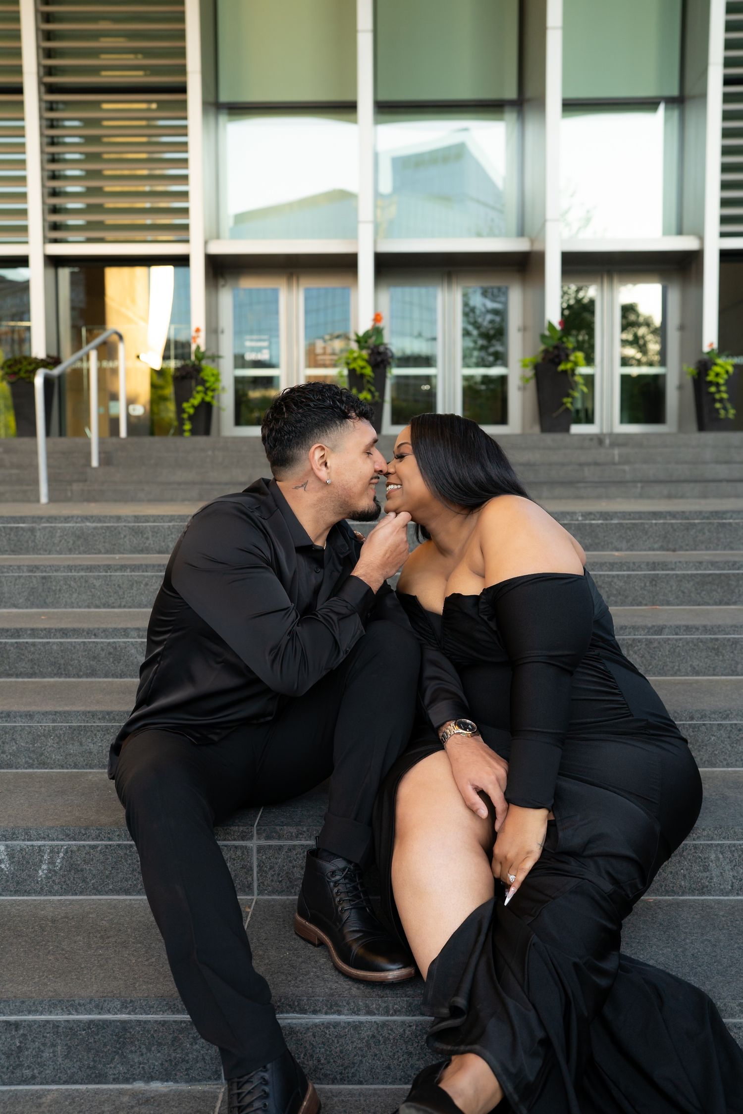 Romantic couple in black formal attire sitting close together on outdoor steps about to kiss.