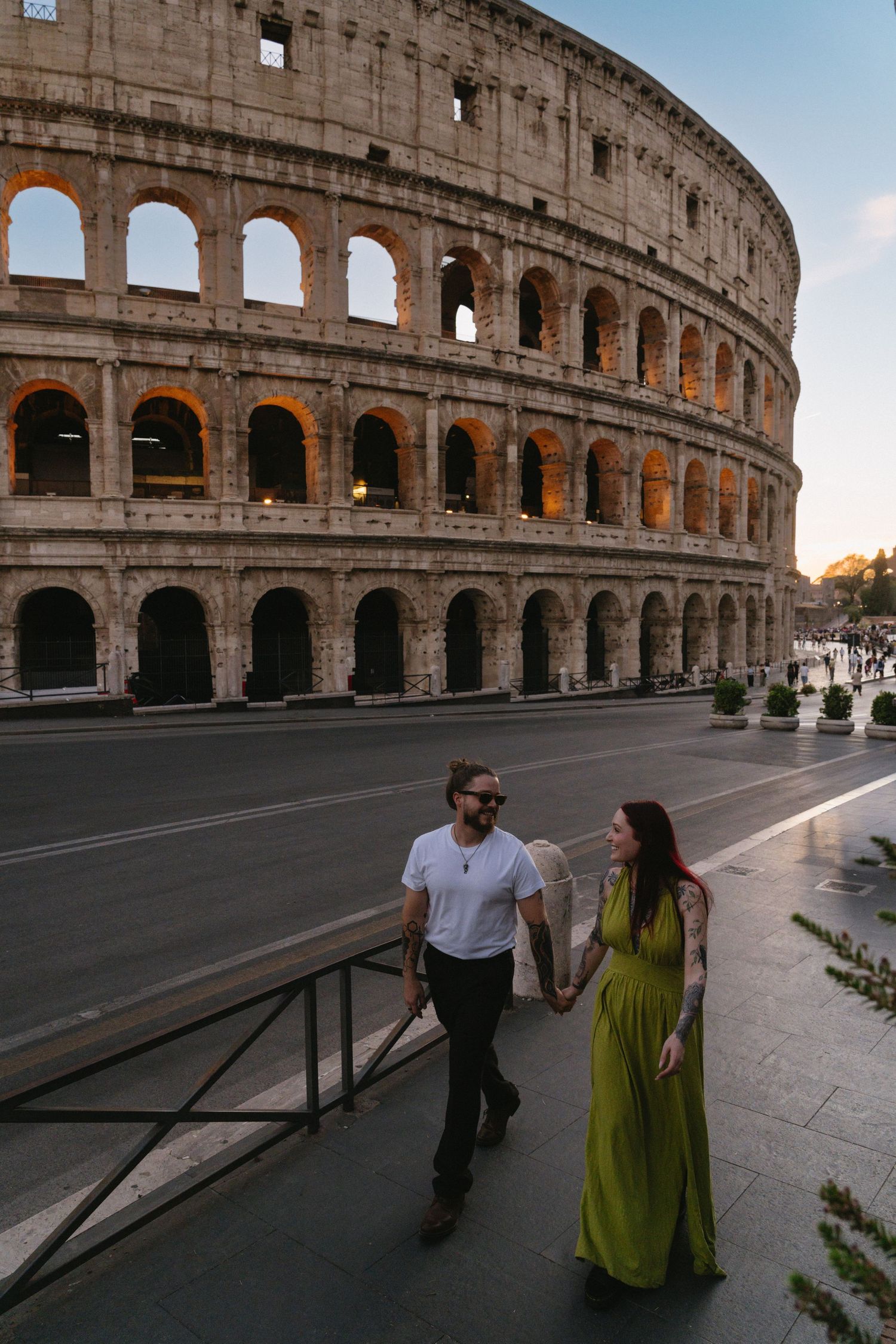 Couple holding hands walking past the illuminated Colosseum in Rome, Italy, at dusk, woman in green dress.