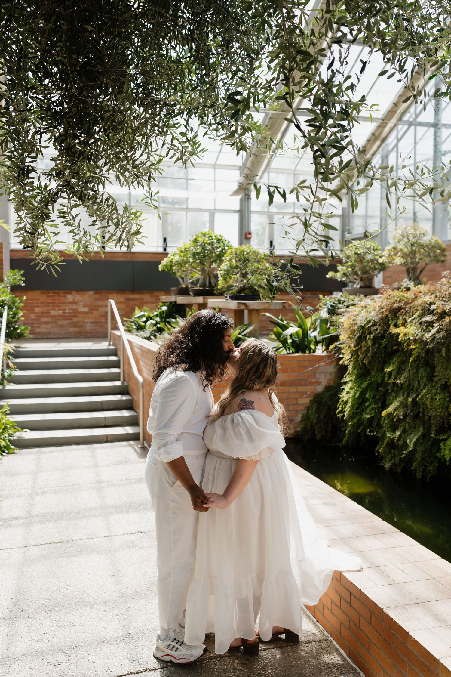 Same-sex couple sharing a kiss in a bright greenhouse garden with brick walls and lush greenery.