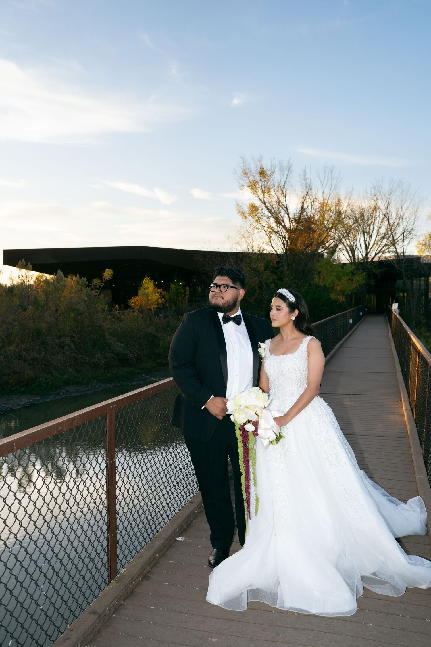 Bride and groom pose on a wooden bridge at sunset surrounded by autumn foliage.