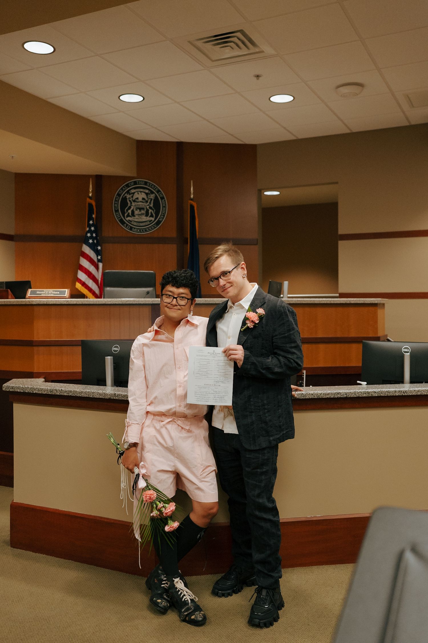 Newlywed couple holding marriage certificate in a courthouse courtroom after wedding ceremony.