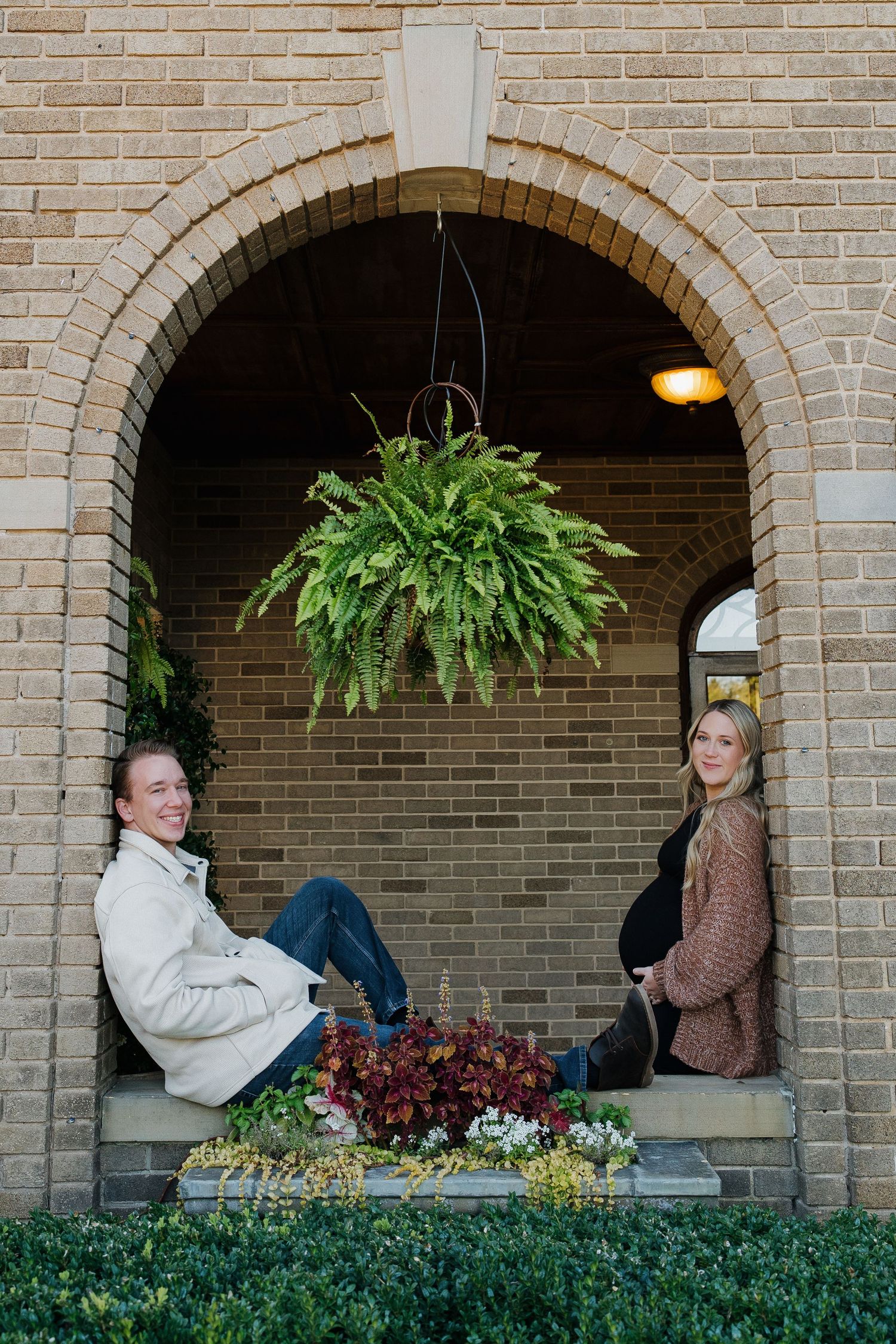 A pregnant couple poses playfully under a brick archway with a lush hanging fern plant.