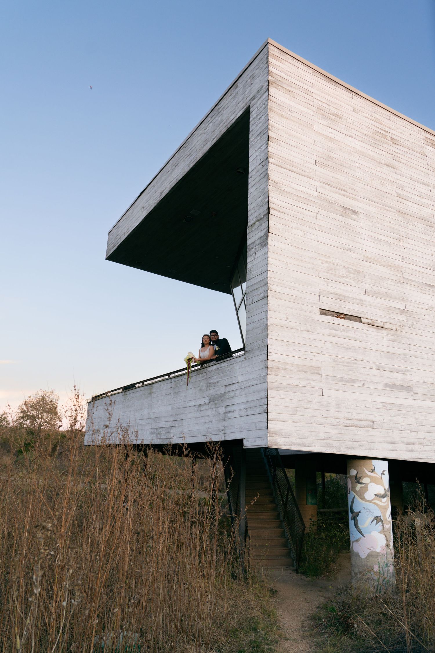 Bride and groom pose on balcony of modern architectural building surrounded by golden field at sunset.