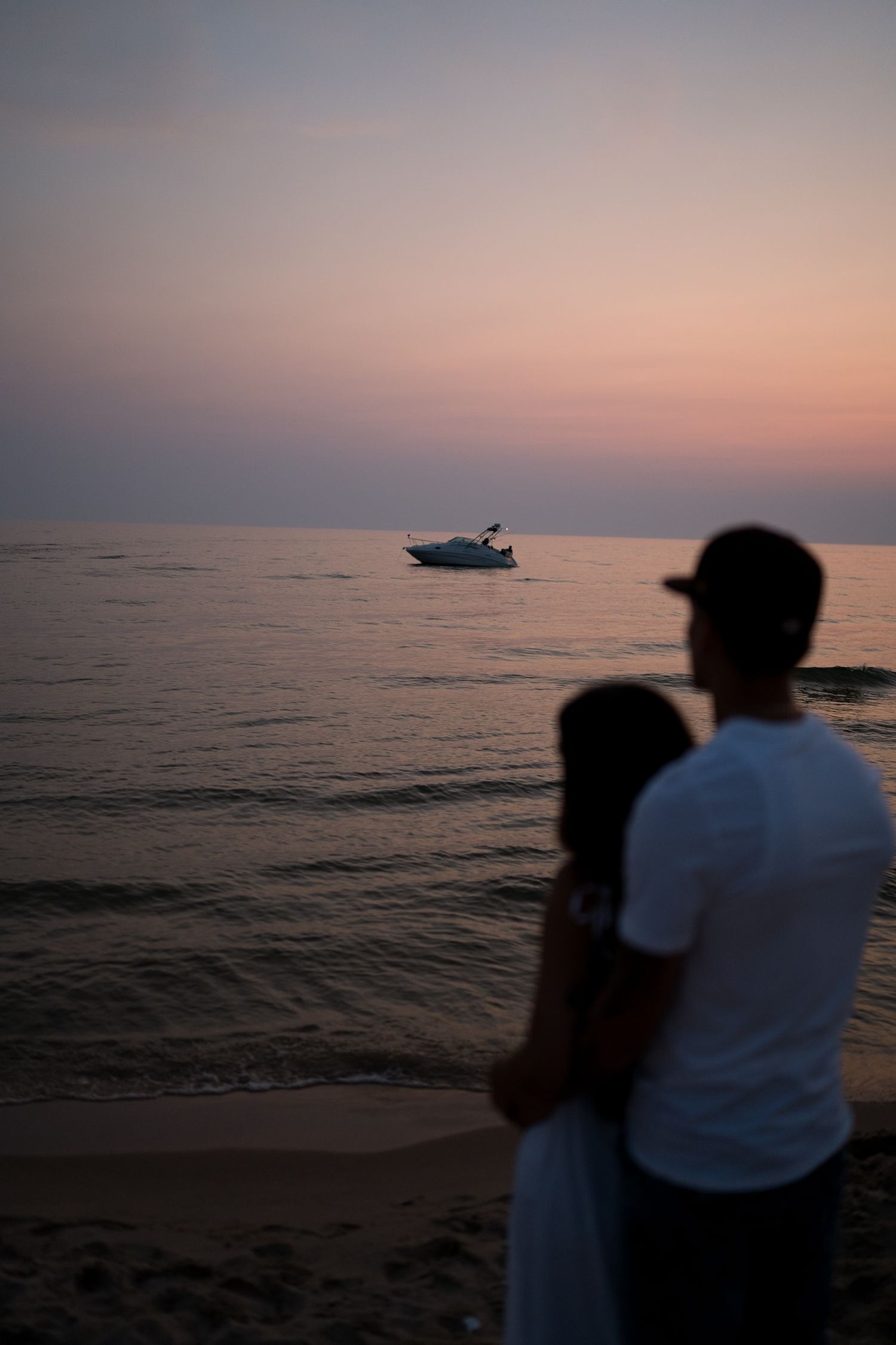 Romantic couple embracing on a sandy beach at sunset watching a boat on calm waters.