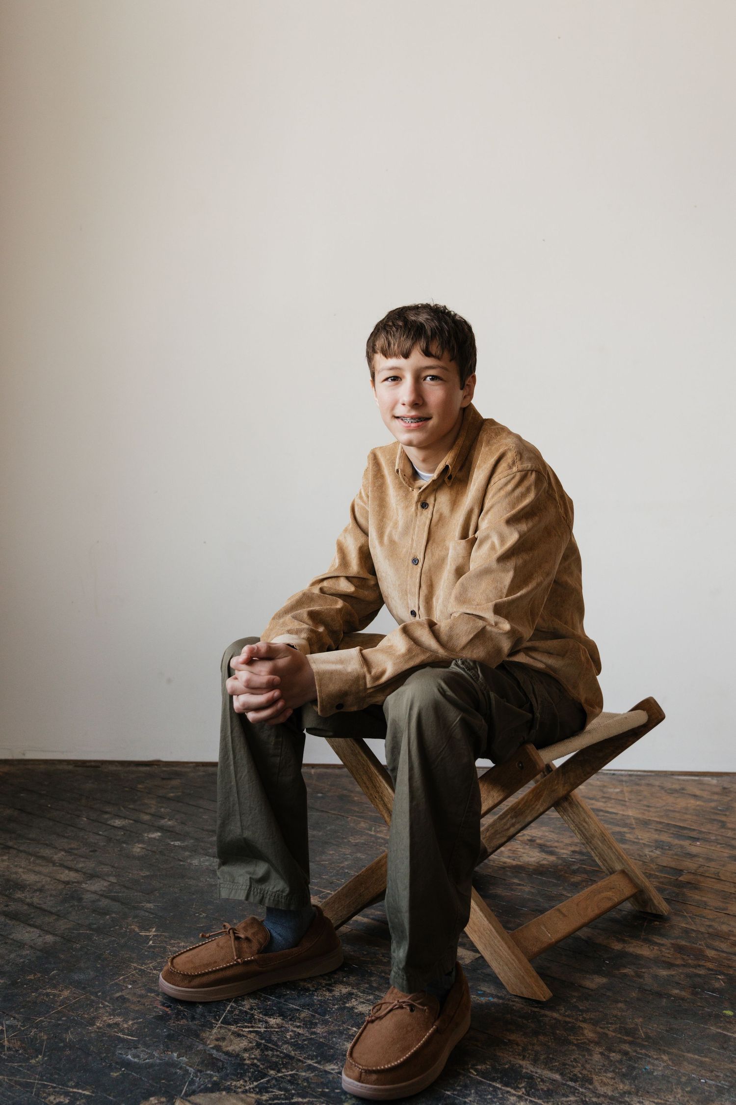 A young man in a tan shirt and olive pants sits relaxed on a wooden stool.