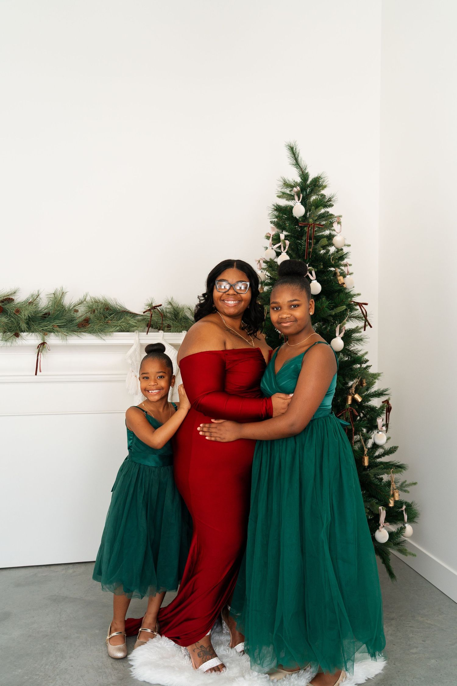 Pregnant mother in red gown smiles with two daughters in green dresses beside a Christmas tree.