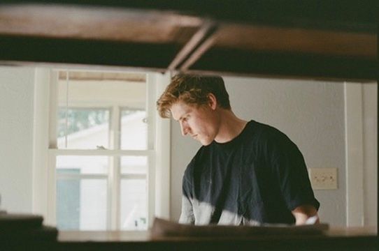 Young man in black t-shirt working at a wooden desk near a bright window in a softly lit room.