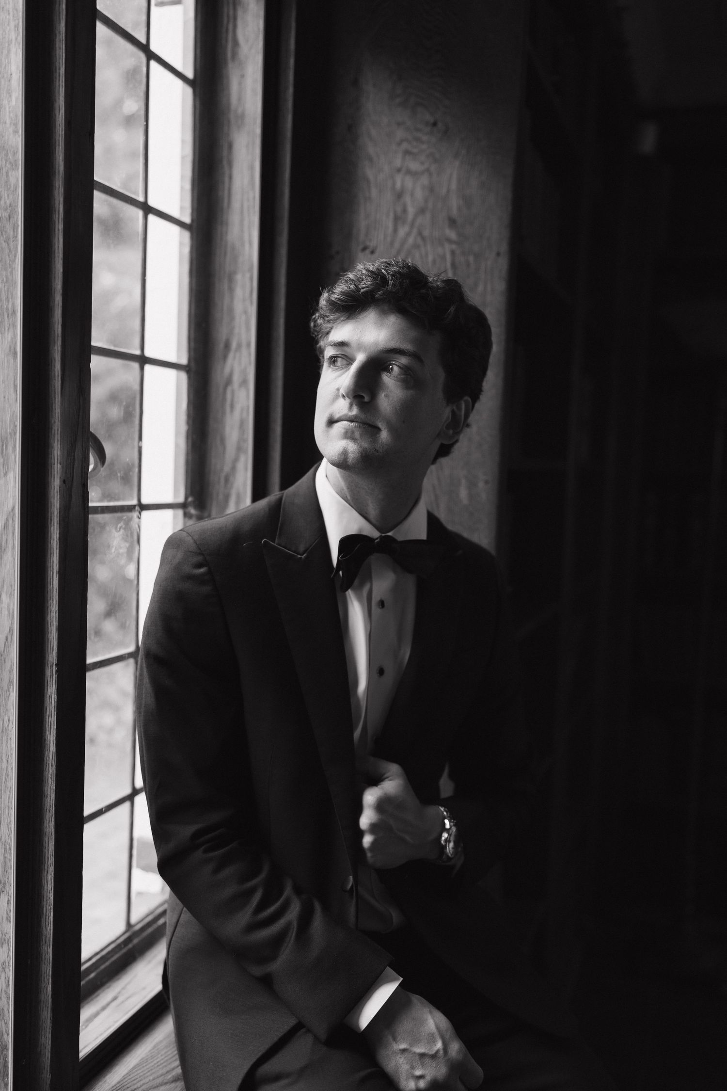 Black and white portrait of groom in tuxedo sitting by leaded glass window looking upward.