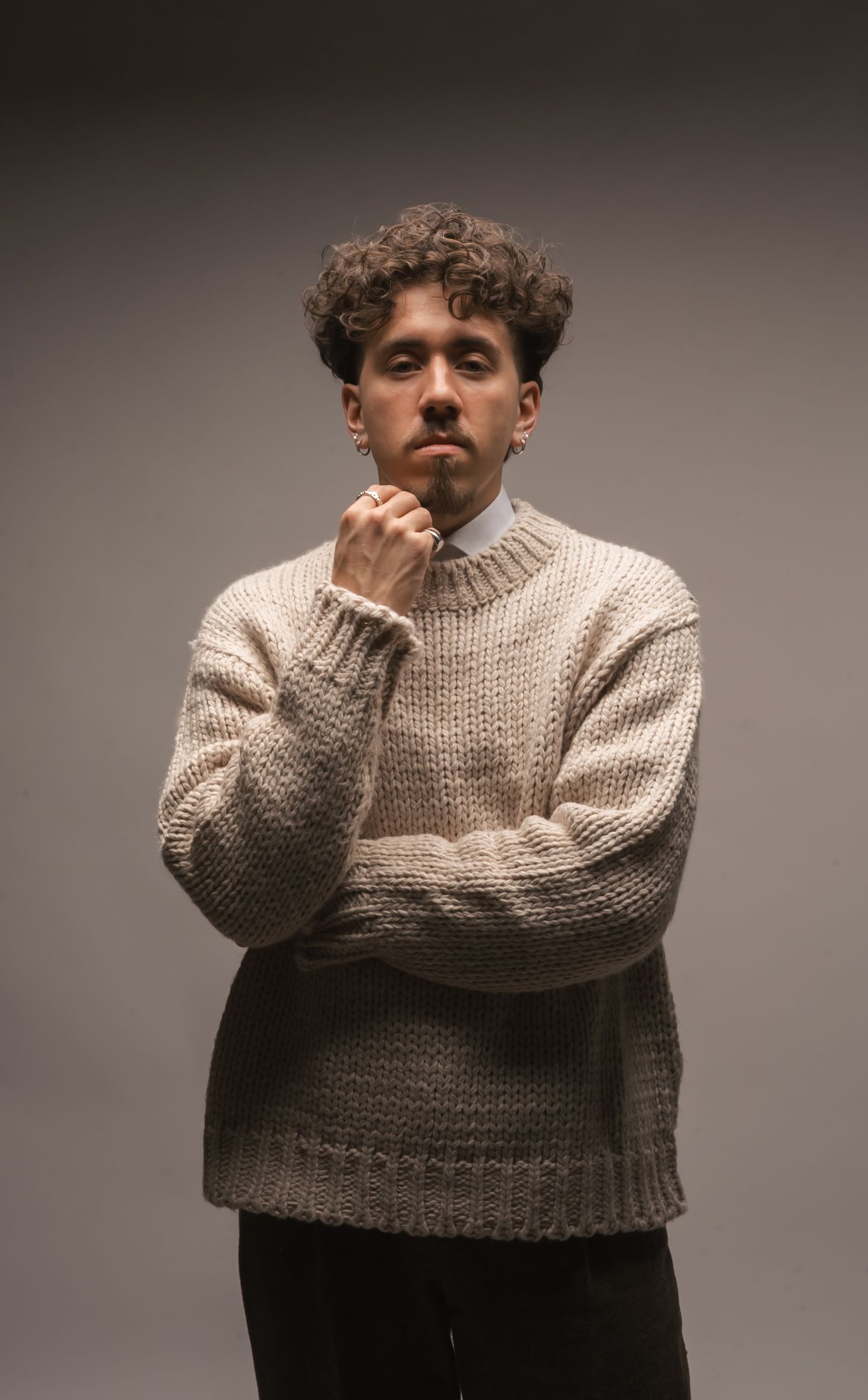 Pensive young man with curly hair wearing a beige chunky knit sweater with hand raised to chin on grey background.