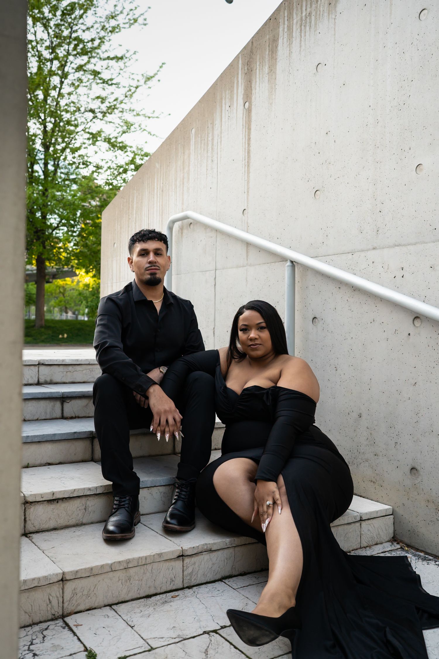 Stylish couple in all-black outfits sitting on outdoor concrete steps near a modern building.
