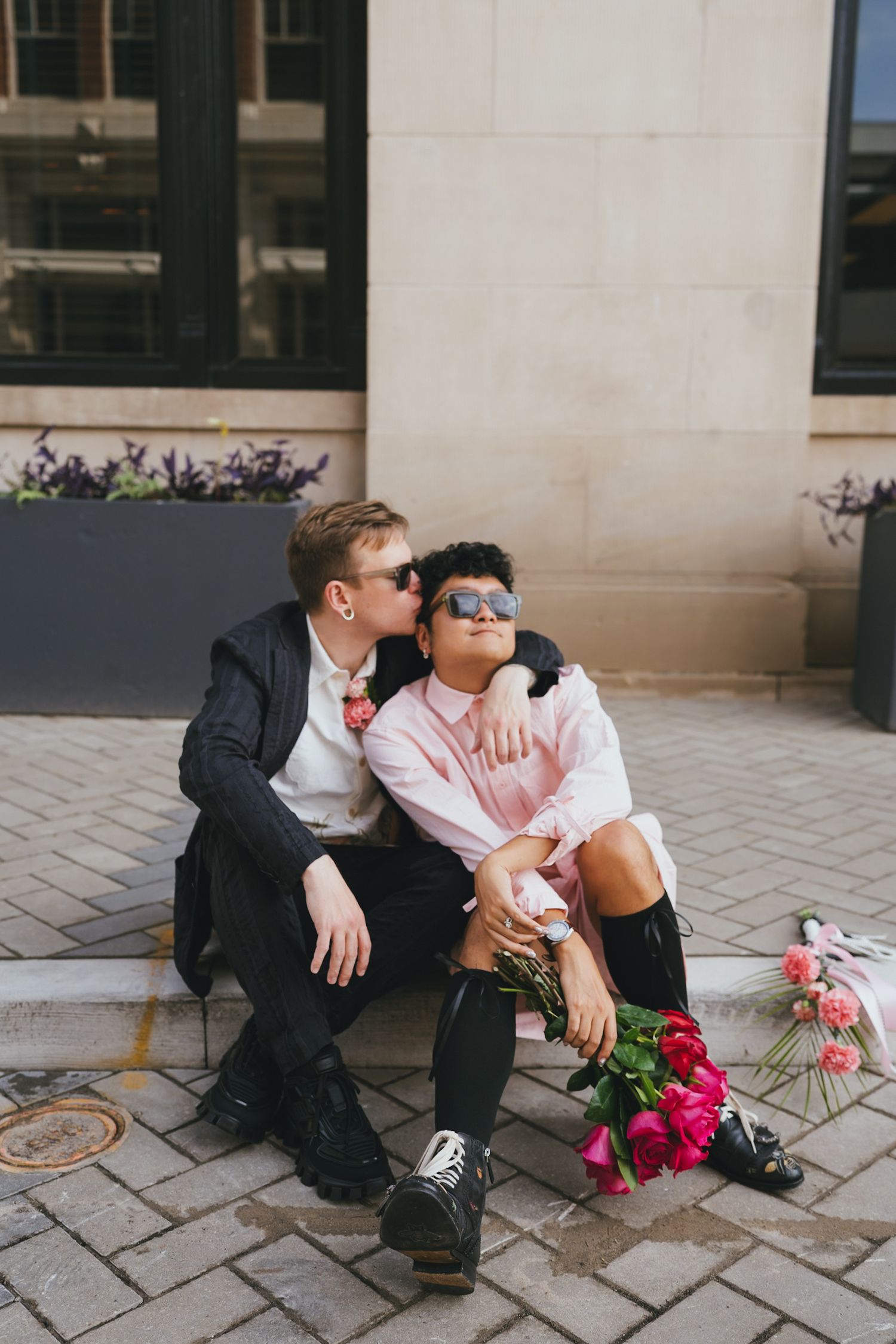 Couple sitting on cobblestone steps, man kissing woman's cheek, holding pink roses, wearing sunglasses and stylish outfits.