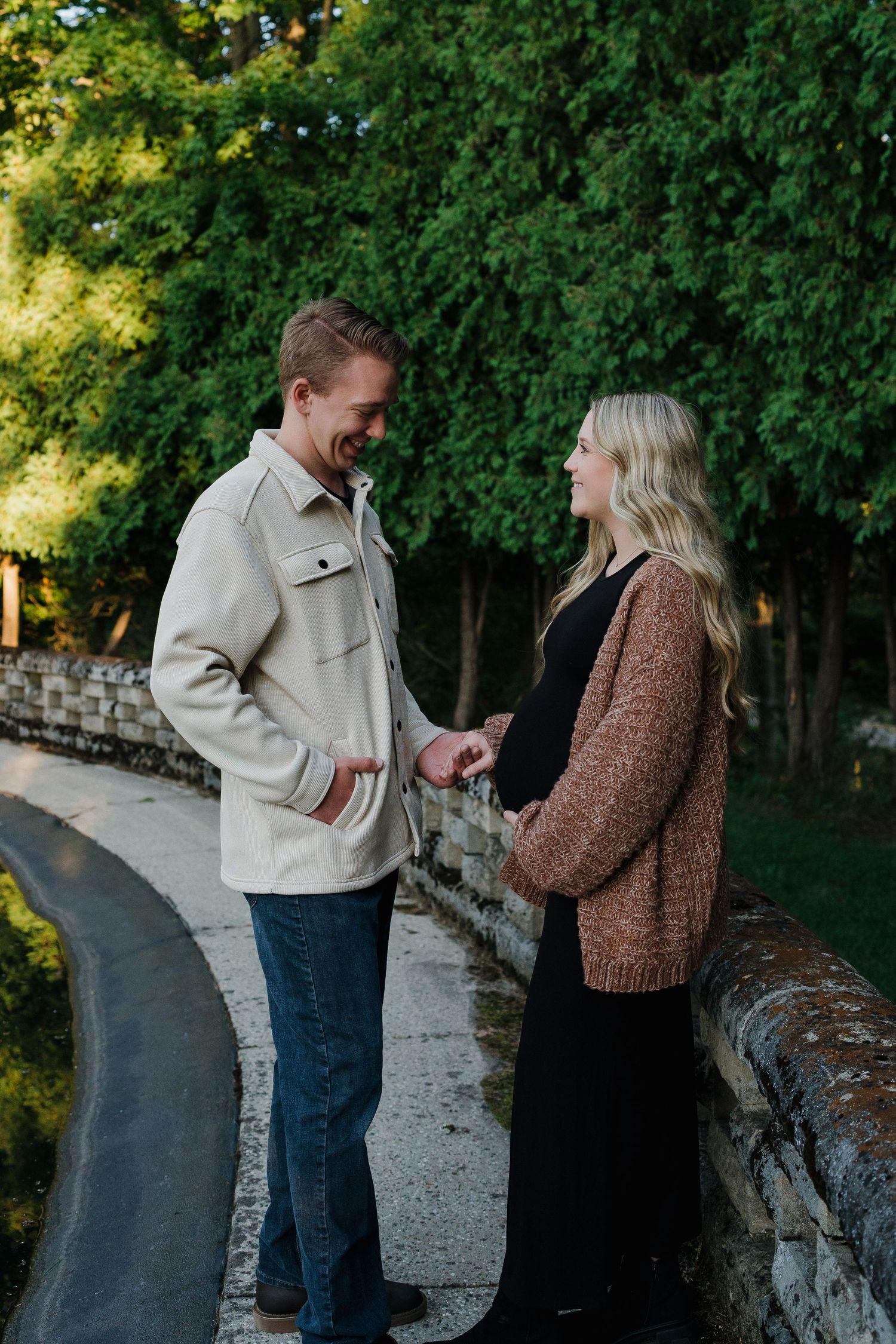 Expectant couple holding hands and smiling at each other on a scenic stone bridge.