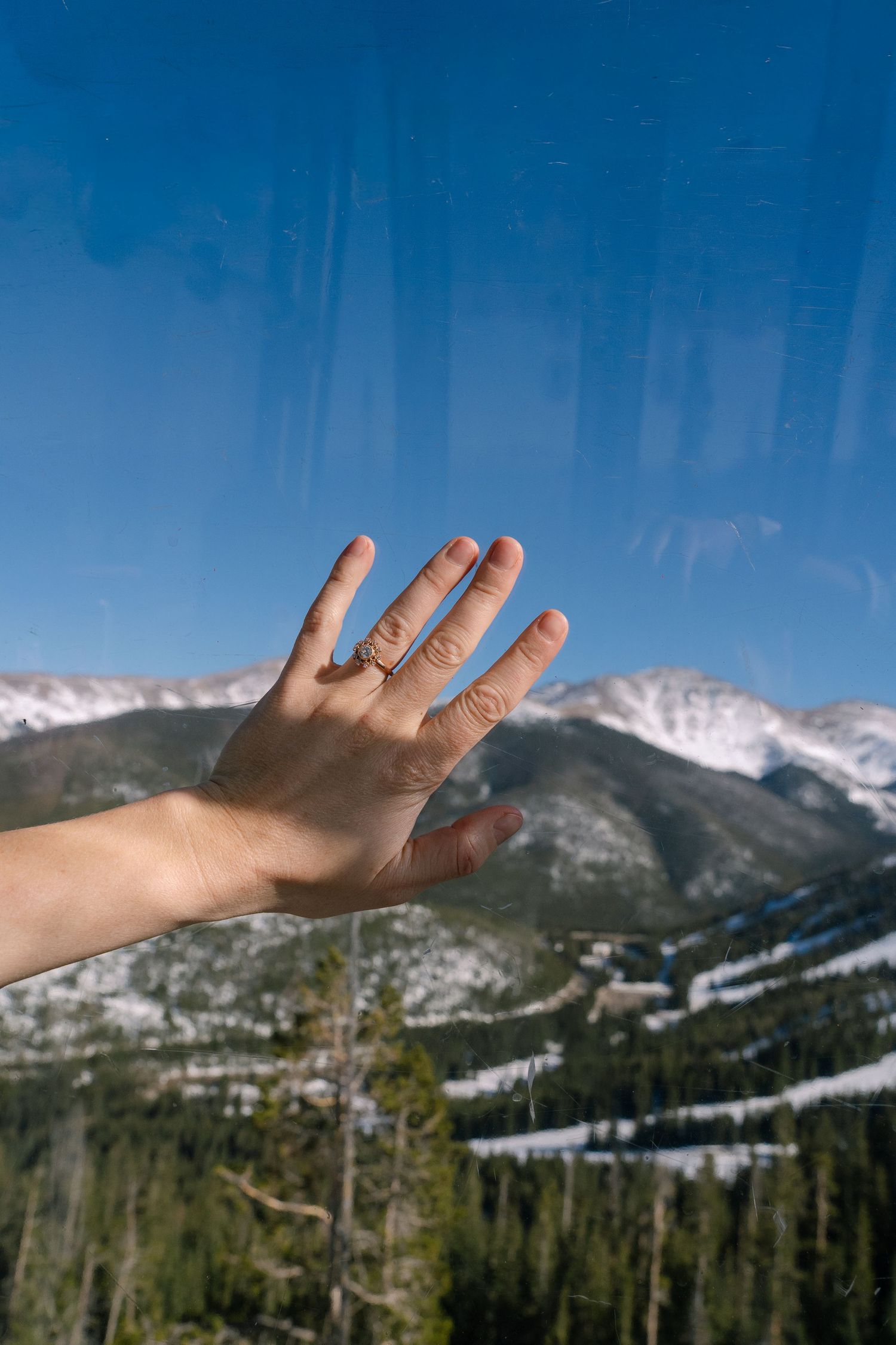 A hand with an engagement ring pressed against a window with snow-capped mountains and pine trees visible in the background.