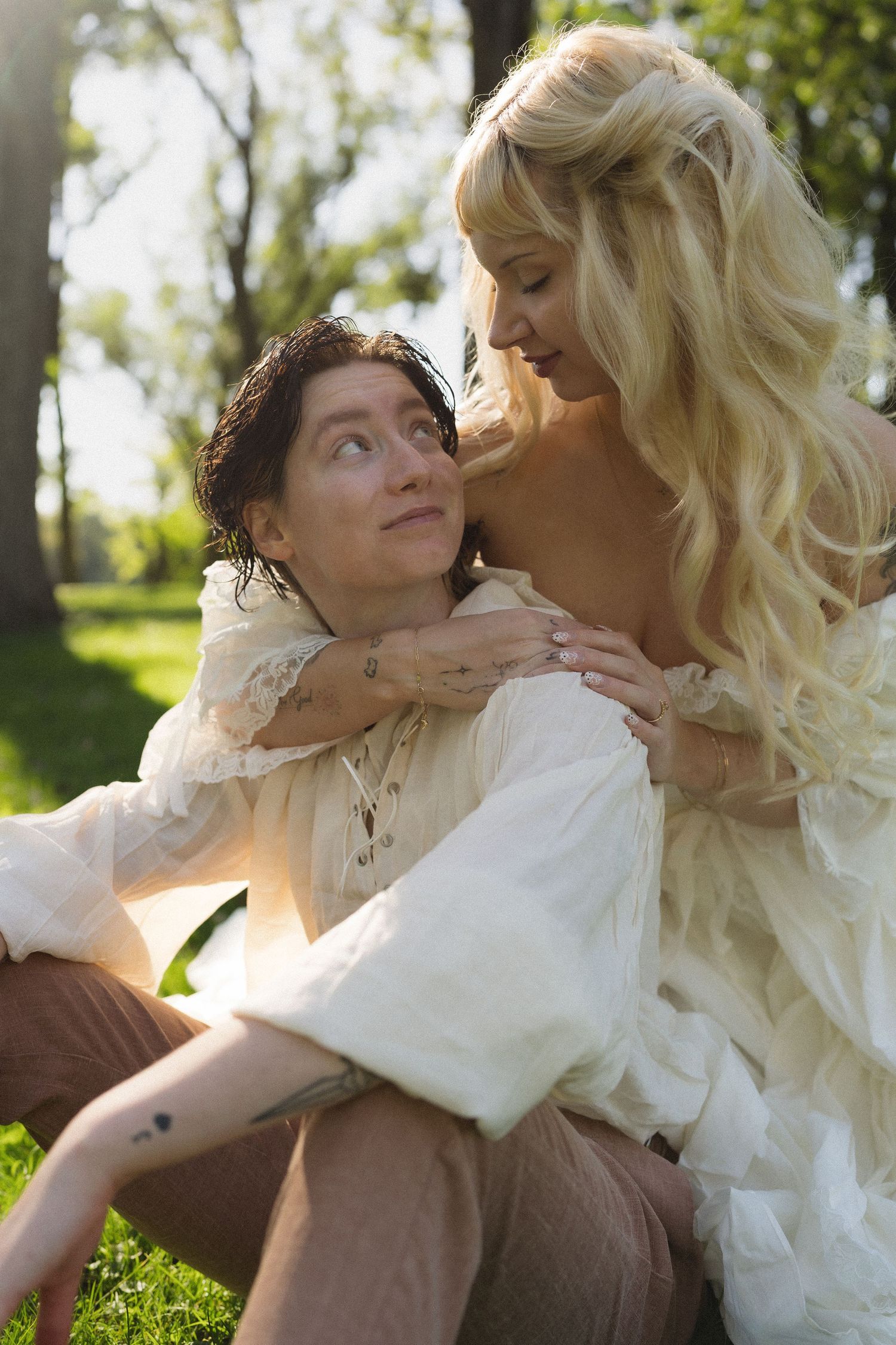 A blonde woman in white lace embraces a dark-haired person sitting on grass in a sunlit park.