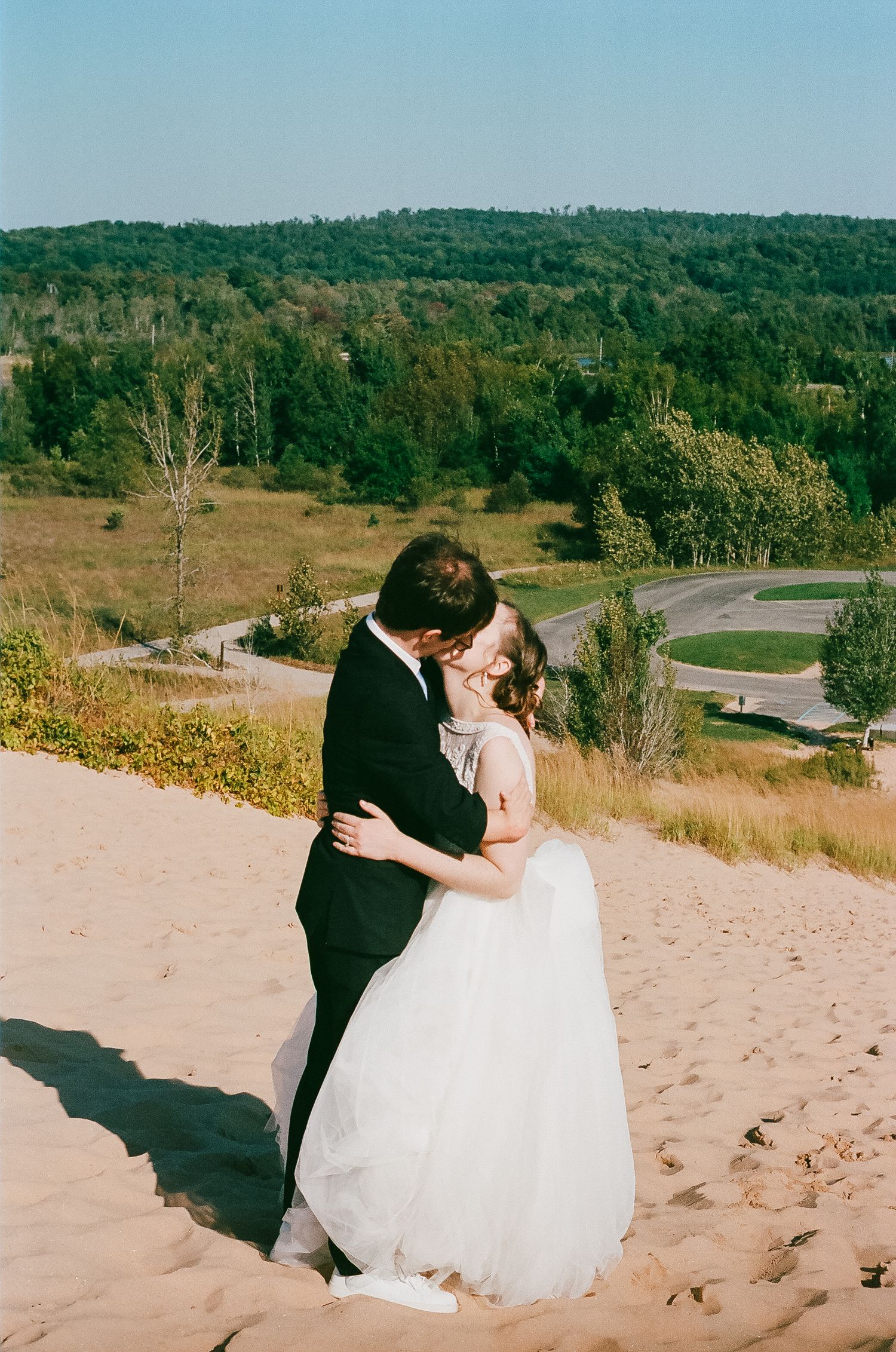 Couple kissing in wedding attire on a sand dune with scenic green valley below.