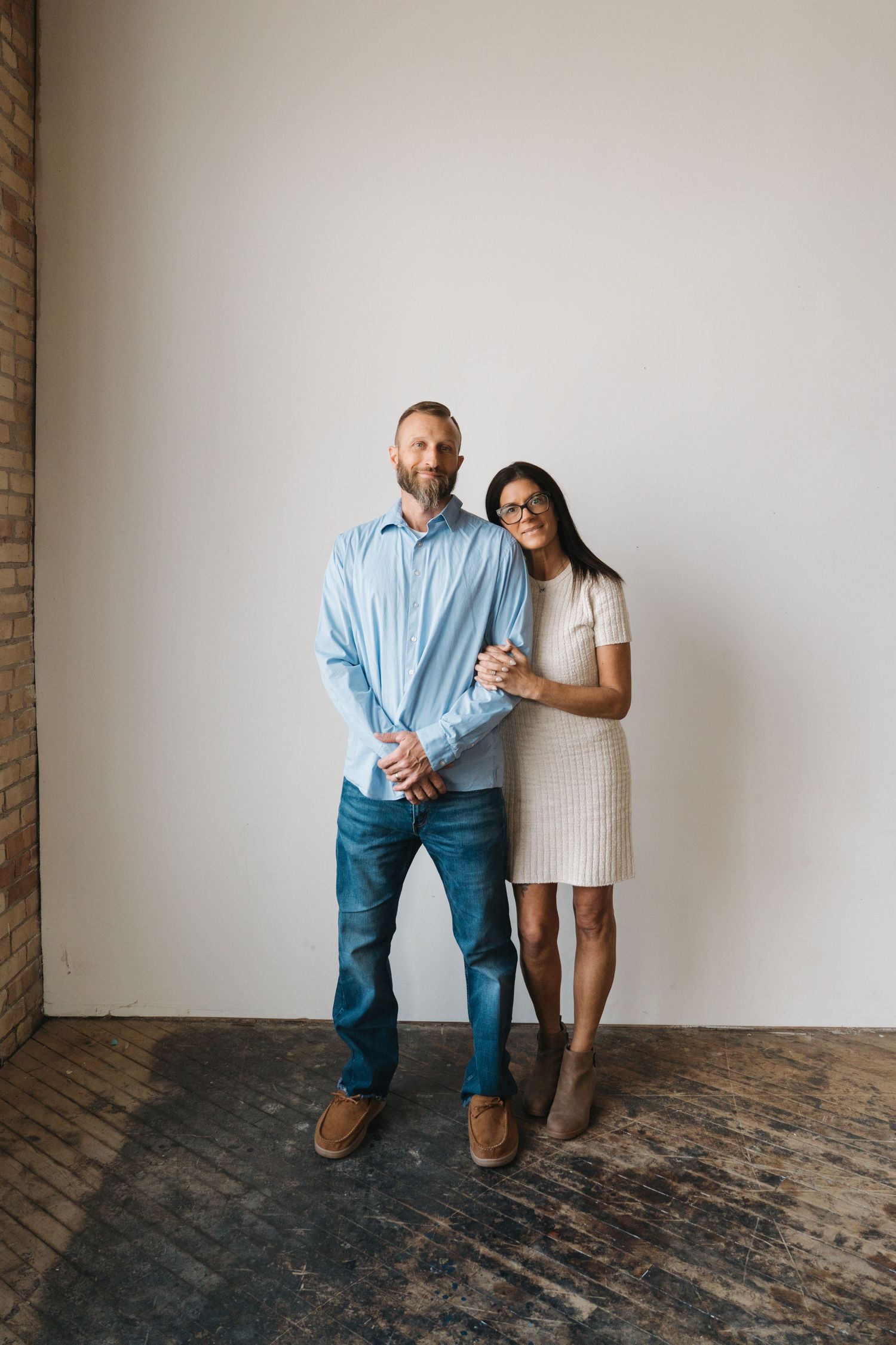Couple posing together in a bright studio, man in blue shirt and jeans, woman in beige knit dress.