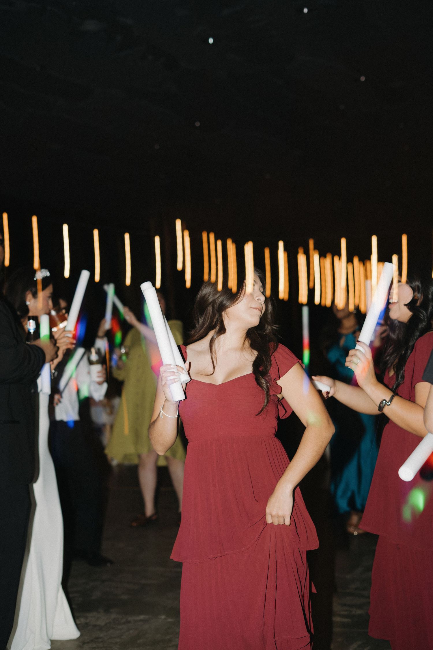 Wedding guest in burgundy dress dancing joyfully with glowing foam sticks at evening reception.