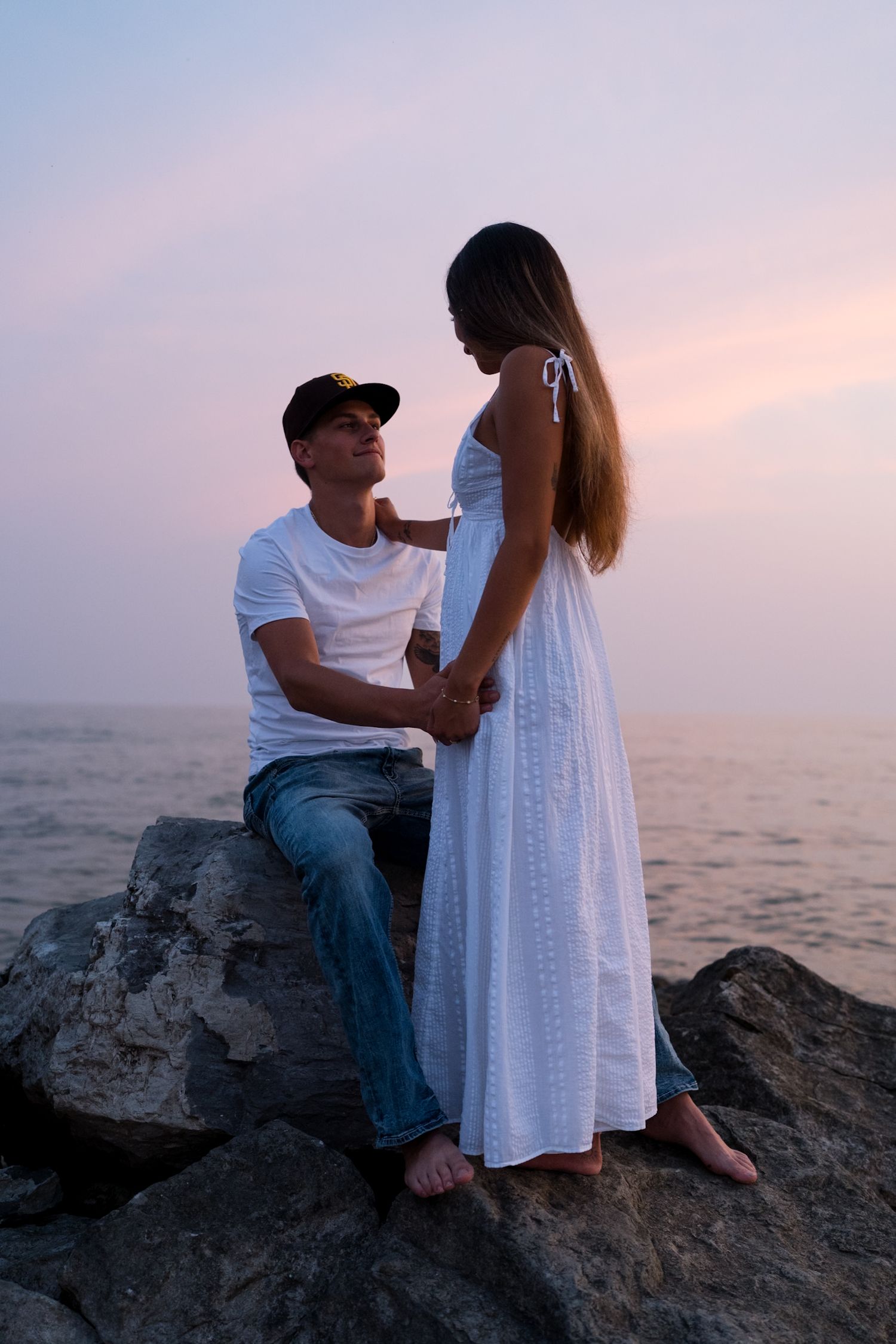Young couple holding hands on rocky beach at sunset, man seated on rocks looking up at woman in white dress.