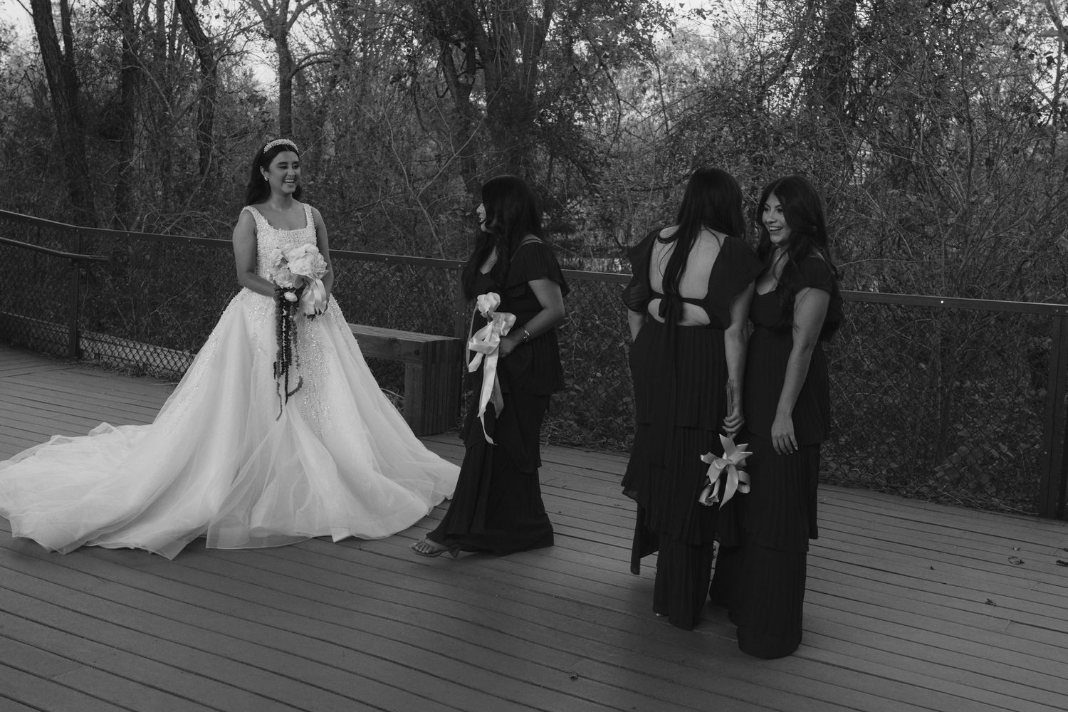 Bride in white gown stands with bridesmaids in black dresses on wooden deck surrounded by trees.