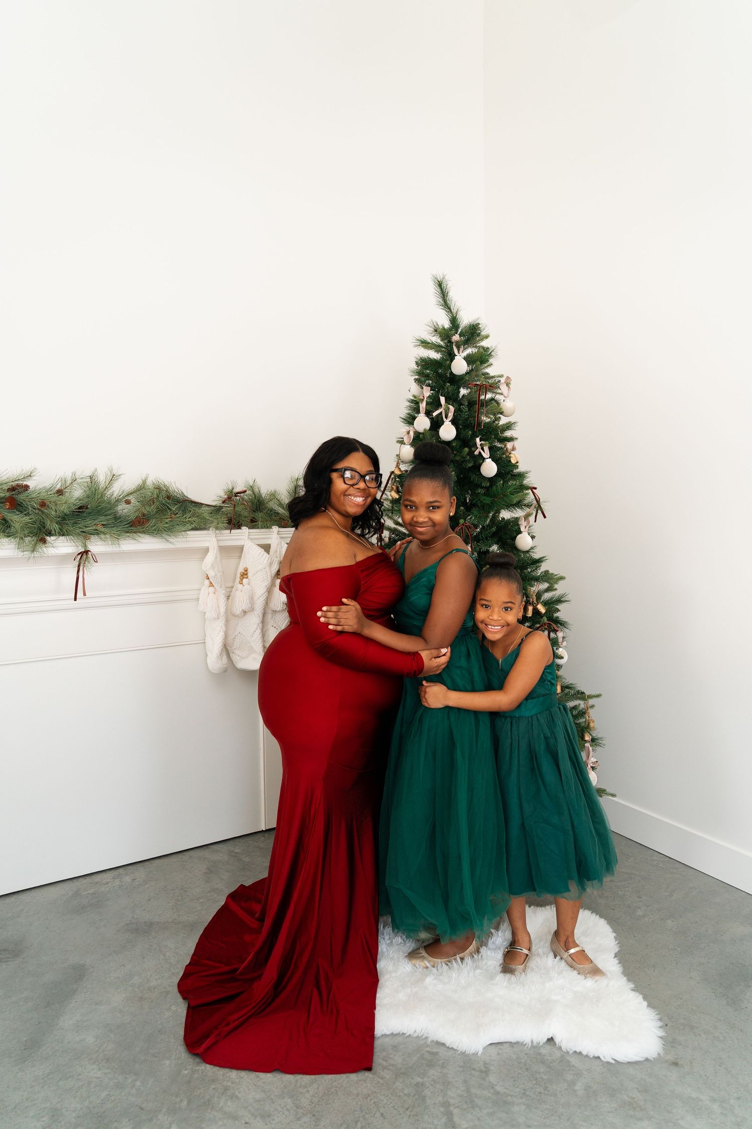 Mother in red maternity gown embraces two daughters in green dresses near a decorated Christmas tree.