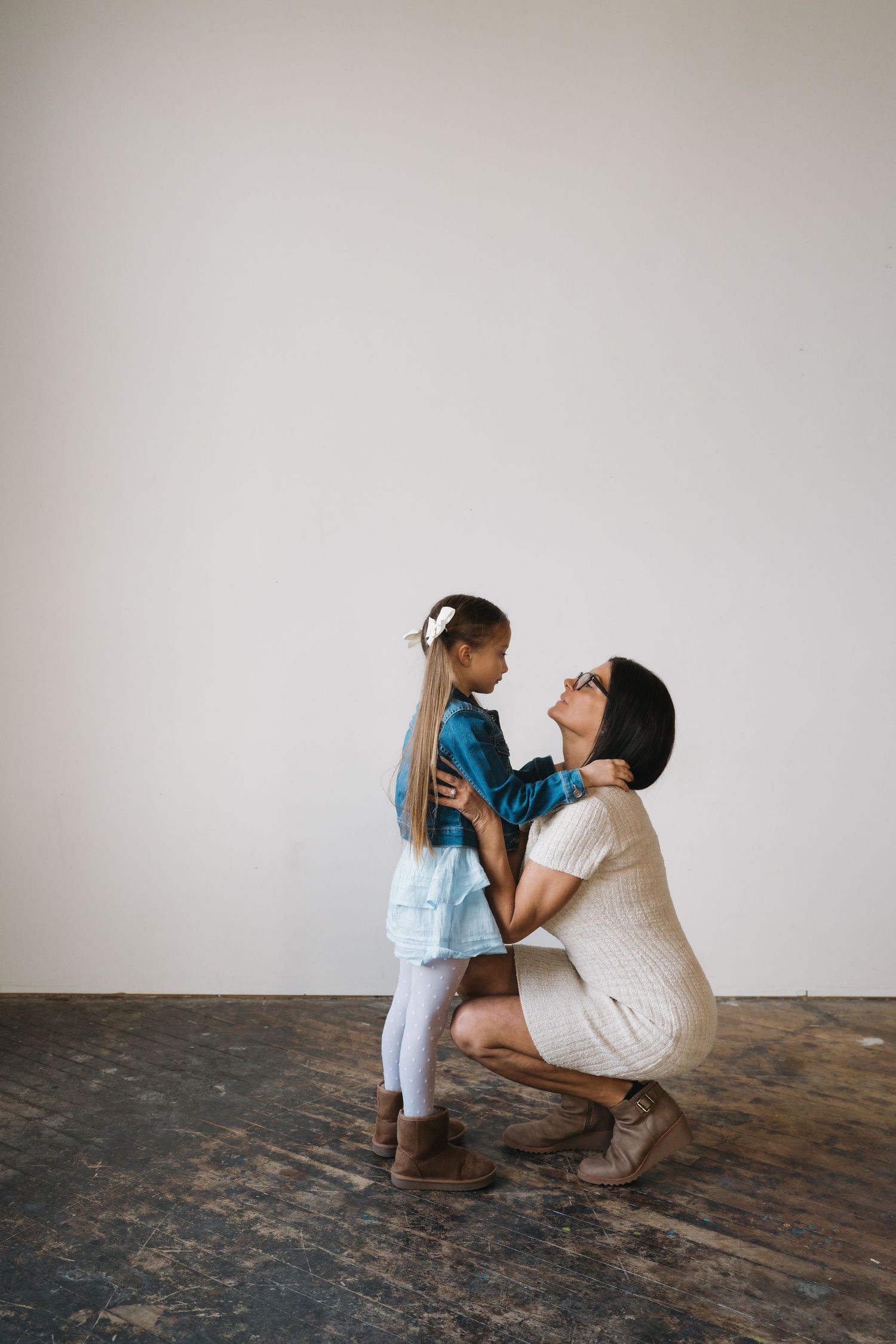 A woman crouches to eye level with a young girl in a blue dress against a white wall.