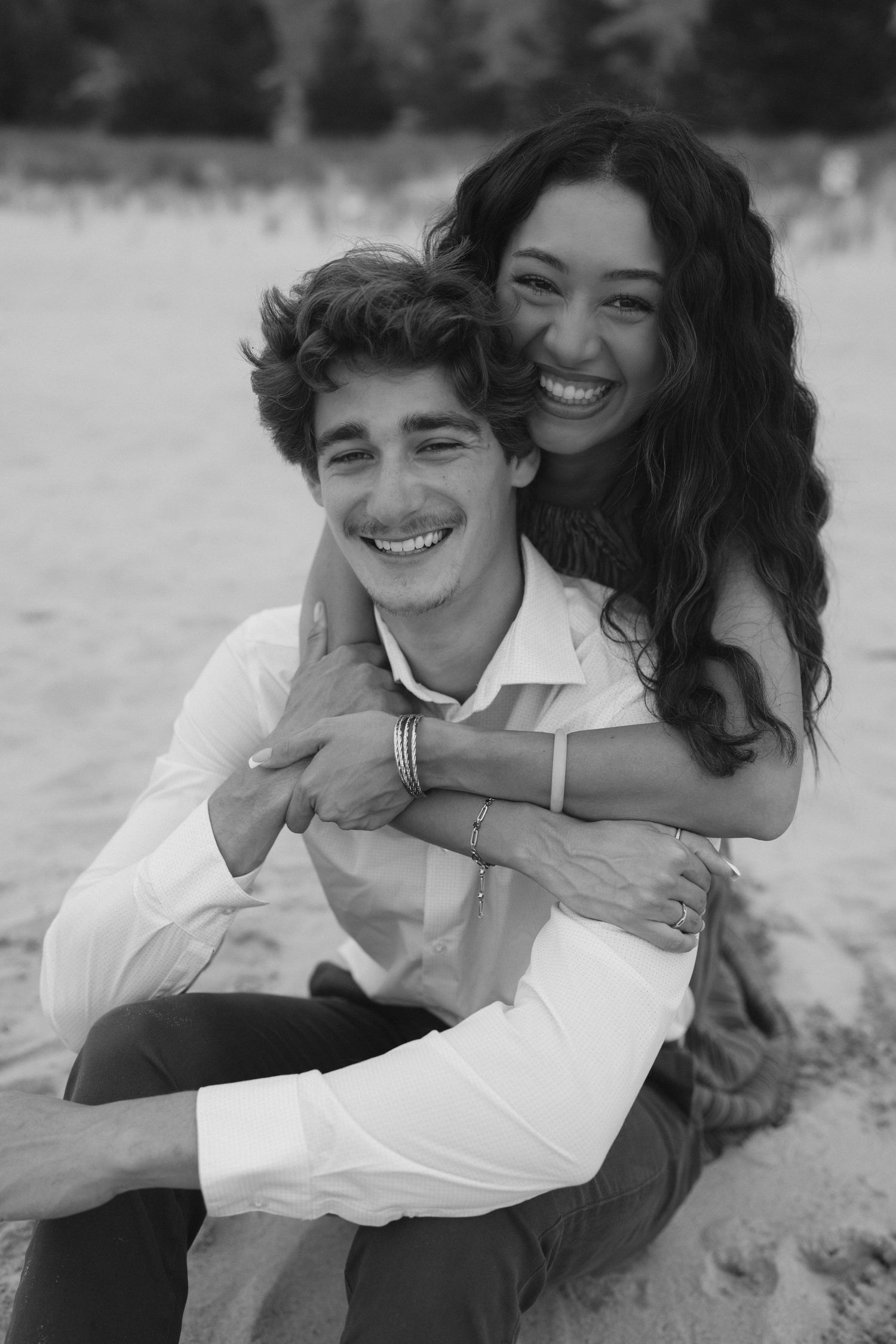 Black and white photo of a smiling couple on a beach, woman hugging man from behind, both laughing joyfully.