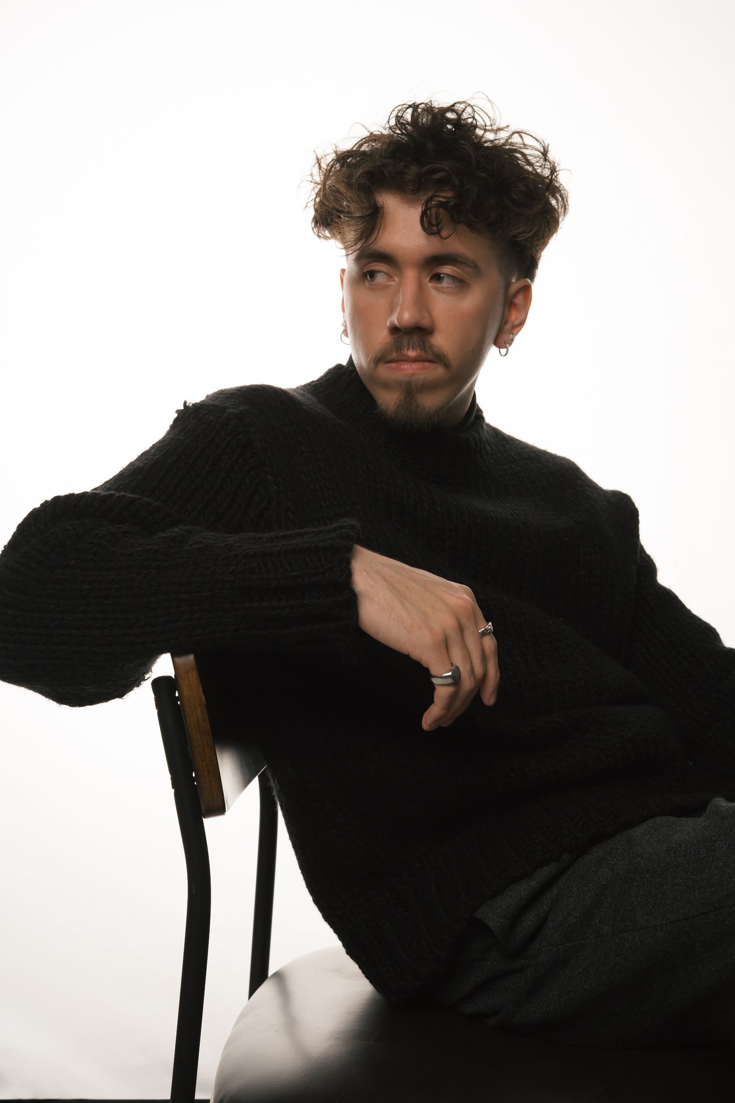 Trendy young man in black turtleneck sweater sitting on chair, looking away thoughtfully with silver ring.