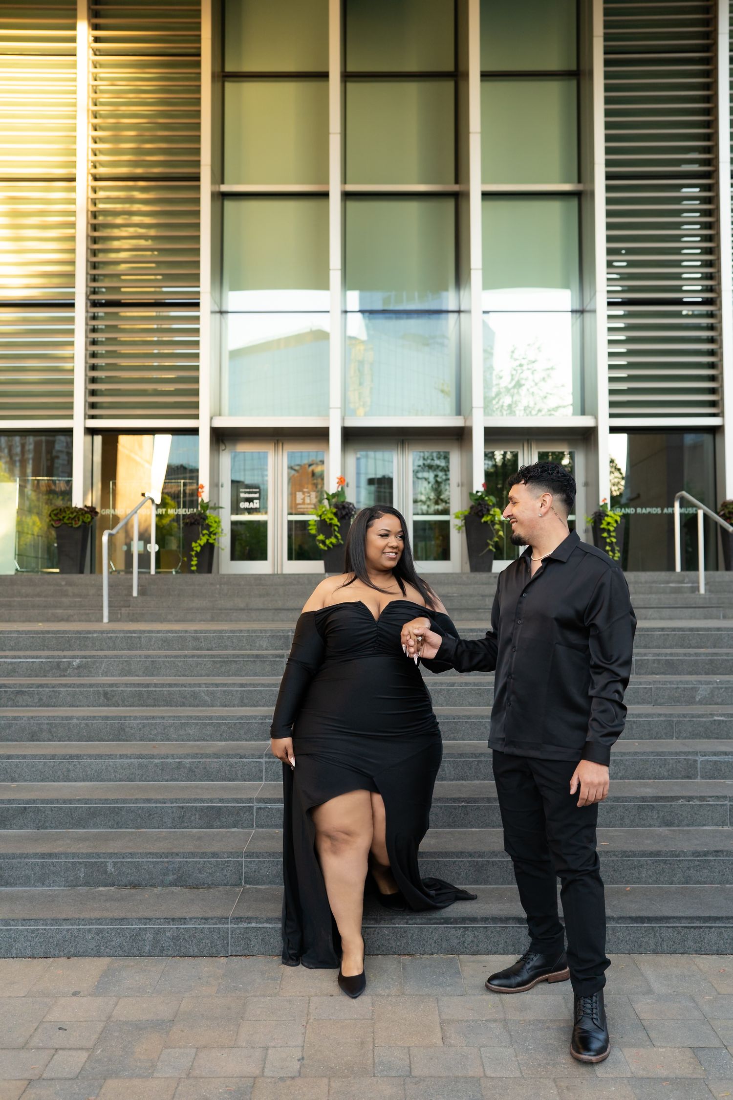 Stylish couple in matching black outfits walking arm-in-arm down steps of a modern building.