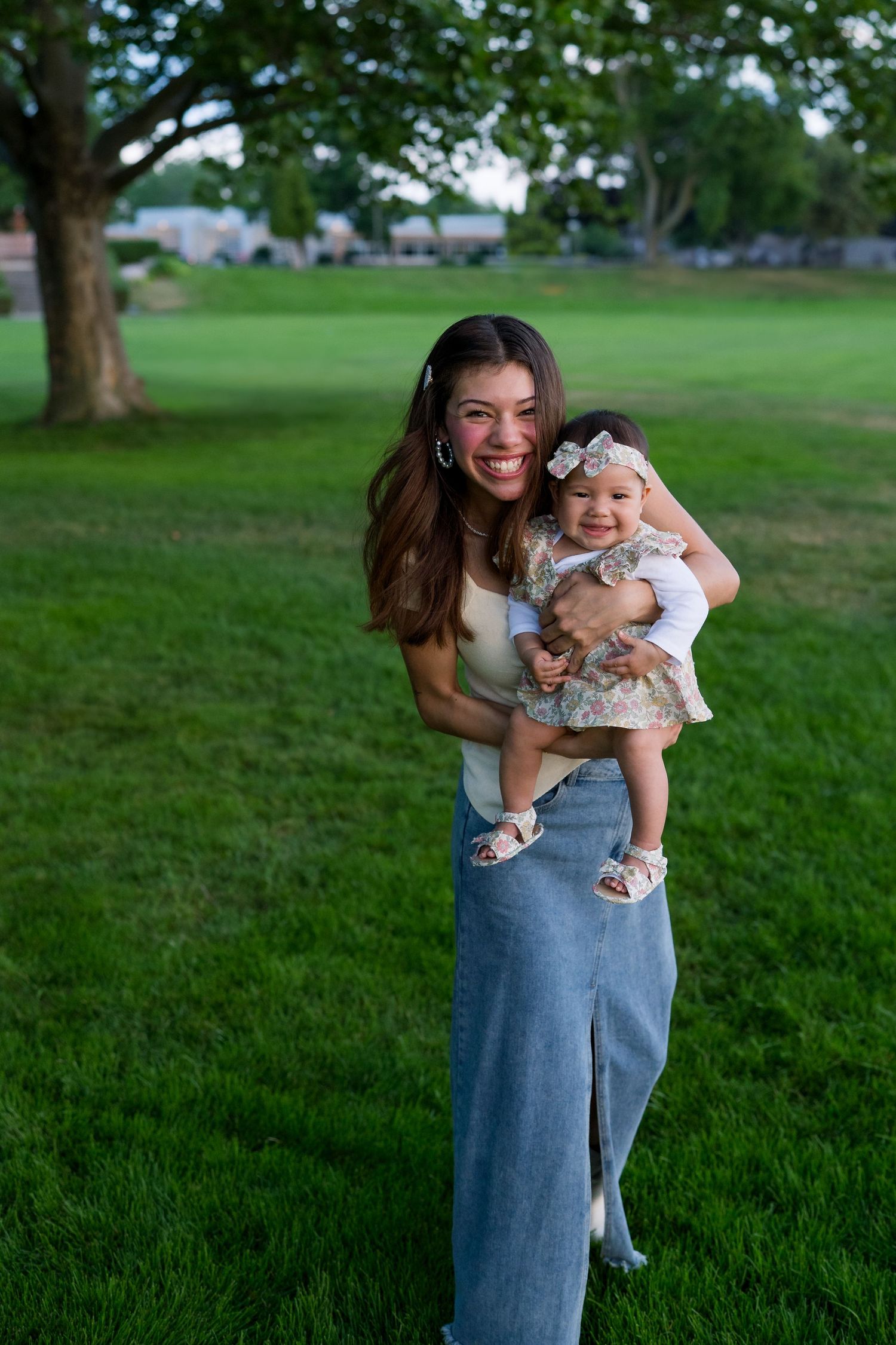 Happy mother standing in green park holding laughing baby girl in floral dress.