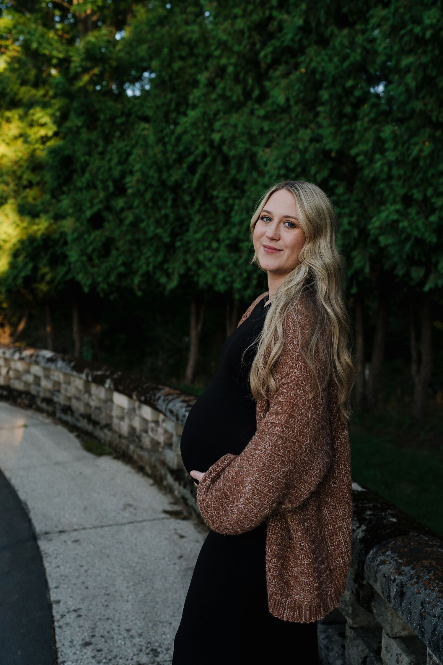 Smiling pregnant woman in black dress and brown cardigan cradling baby bump by a stone wall outdoors.