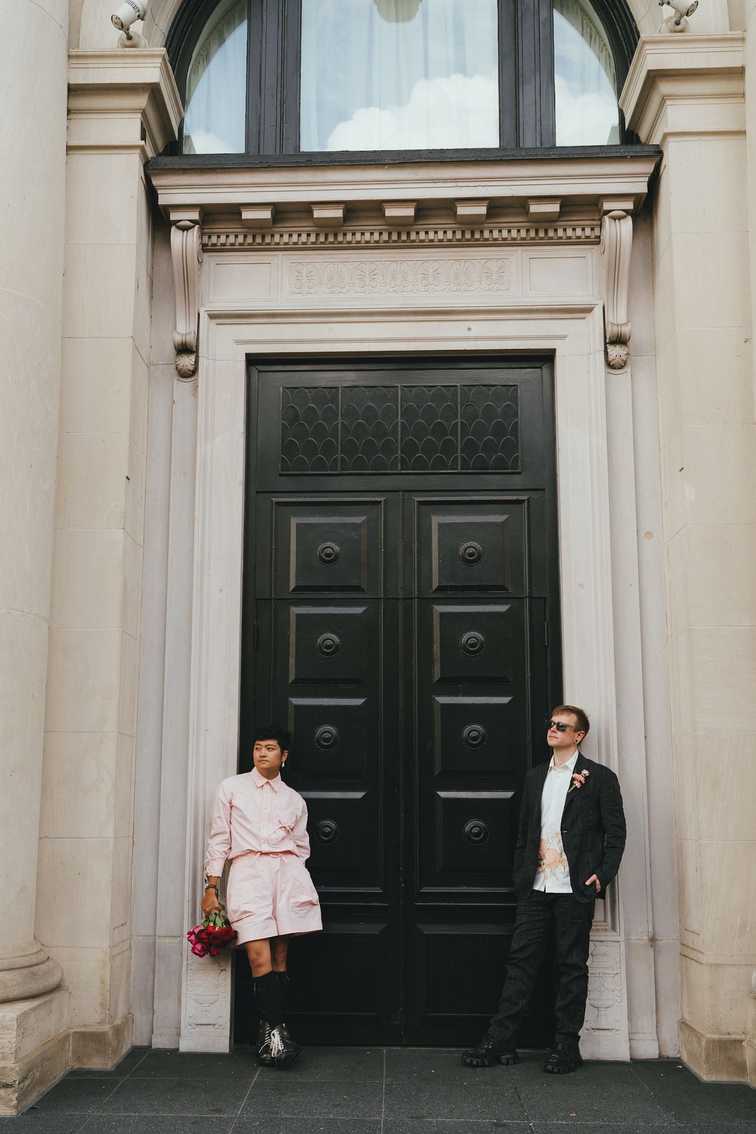 Couple standing before large black ornate door, woman in pink outfit holding flowers, man in dark suit with sunglasses.