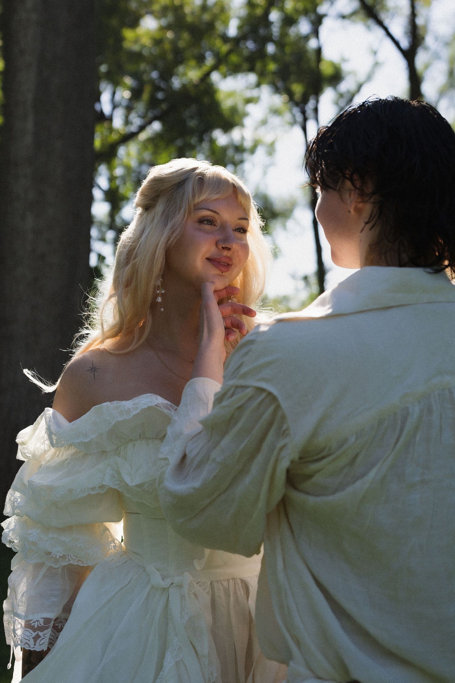 A blonde woman in a white ruffled gown gazes at a dark-haired person outdoors among trees in romantic attire.