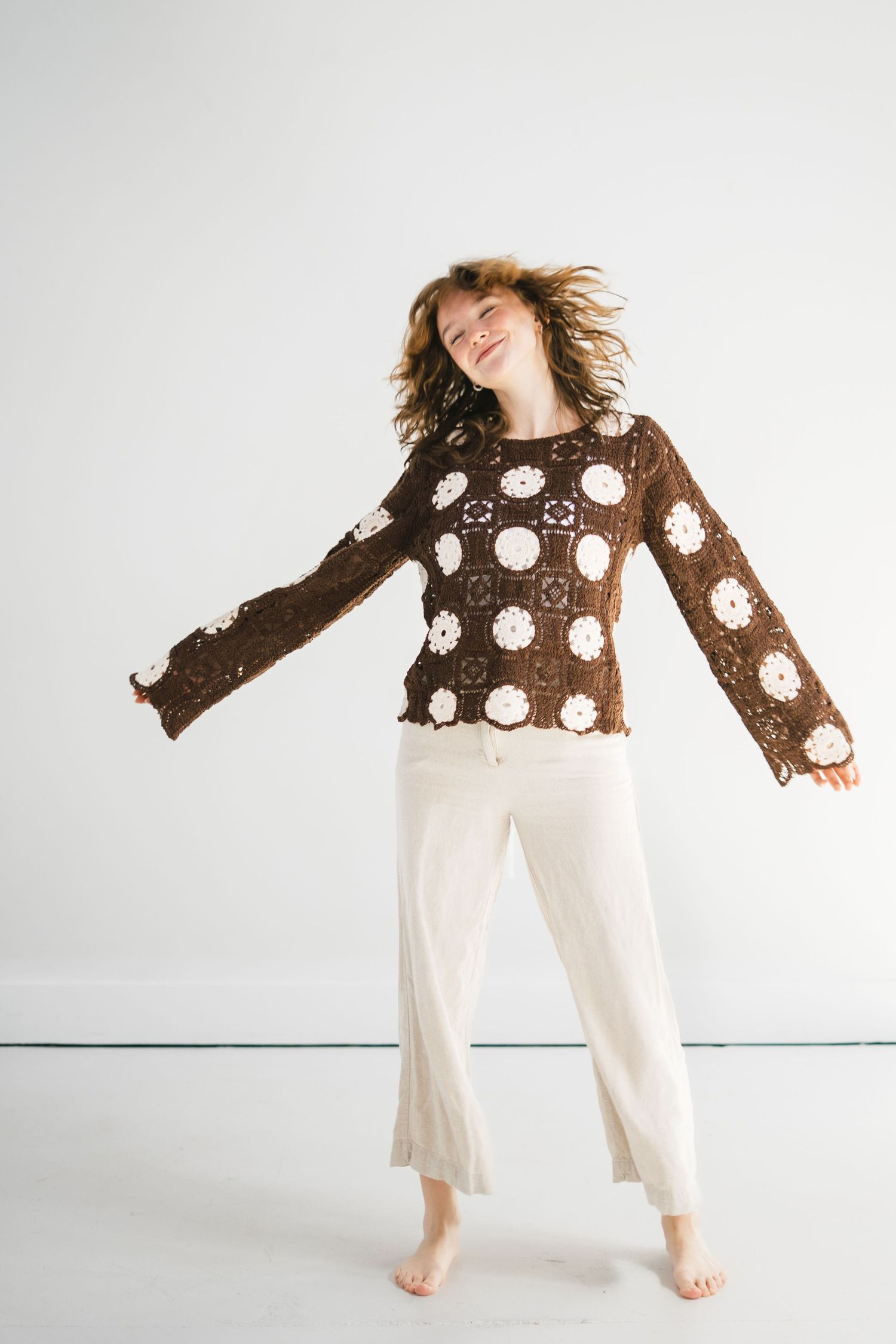 A joyful barefoot woman in a brown crochet floral sweater and white wide-leg pants dances with arms outstretched.