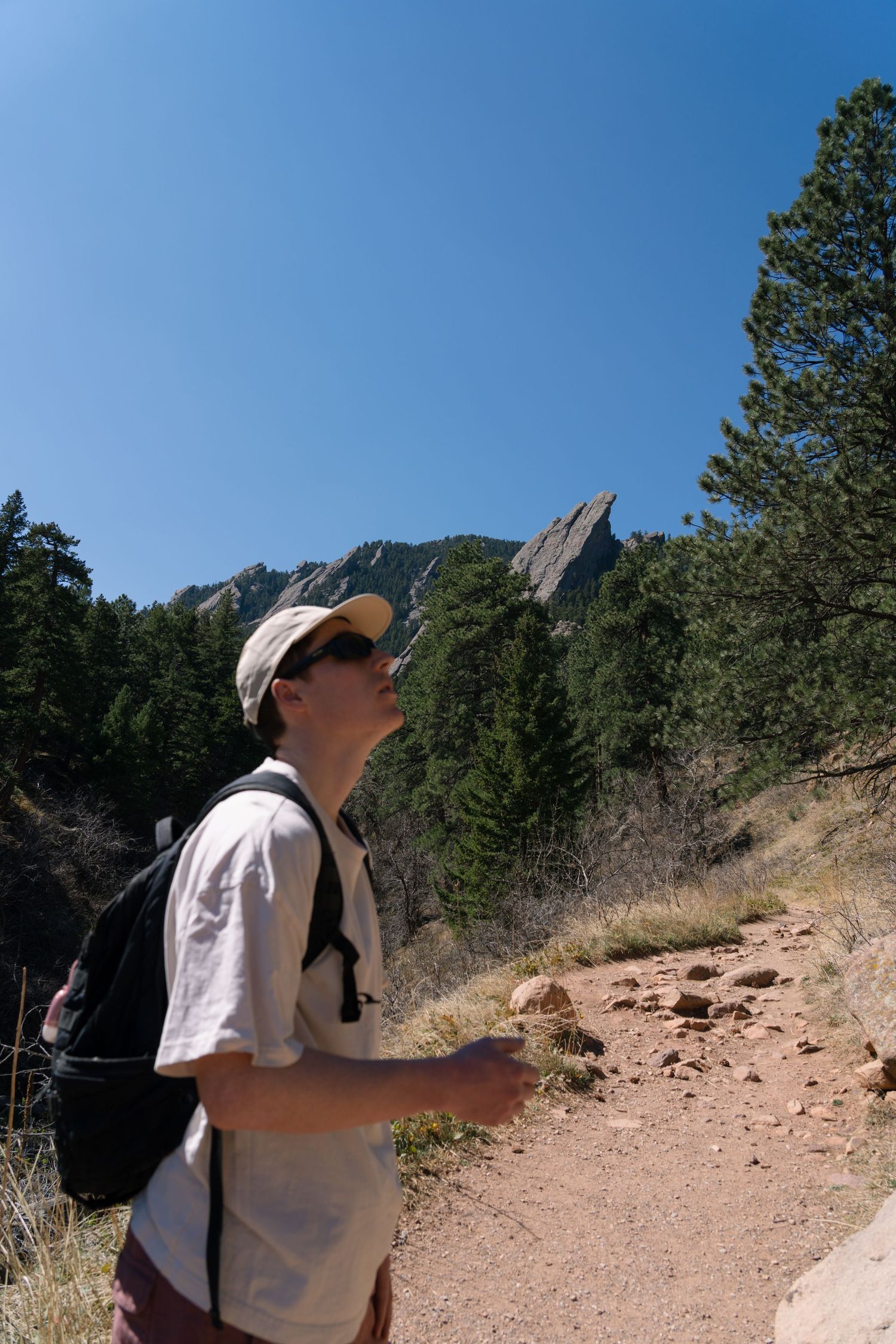 Hiker with backpack and cap looks up at the Flatirons rock formations along a Colorado trail.