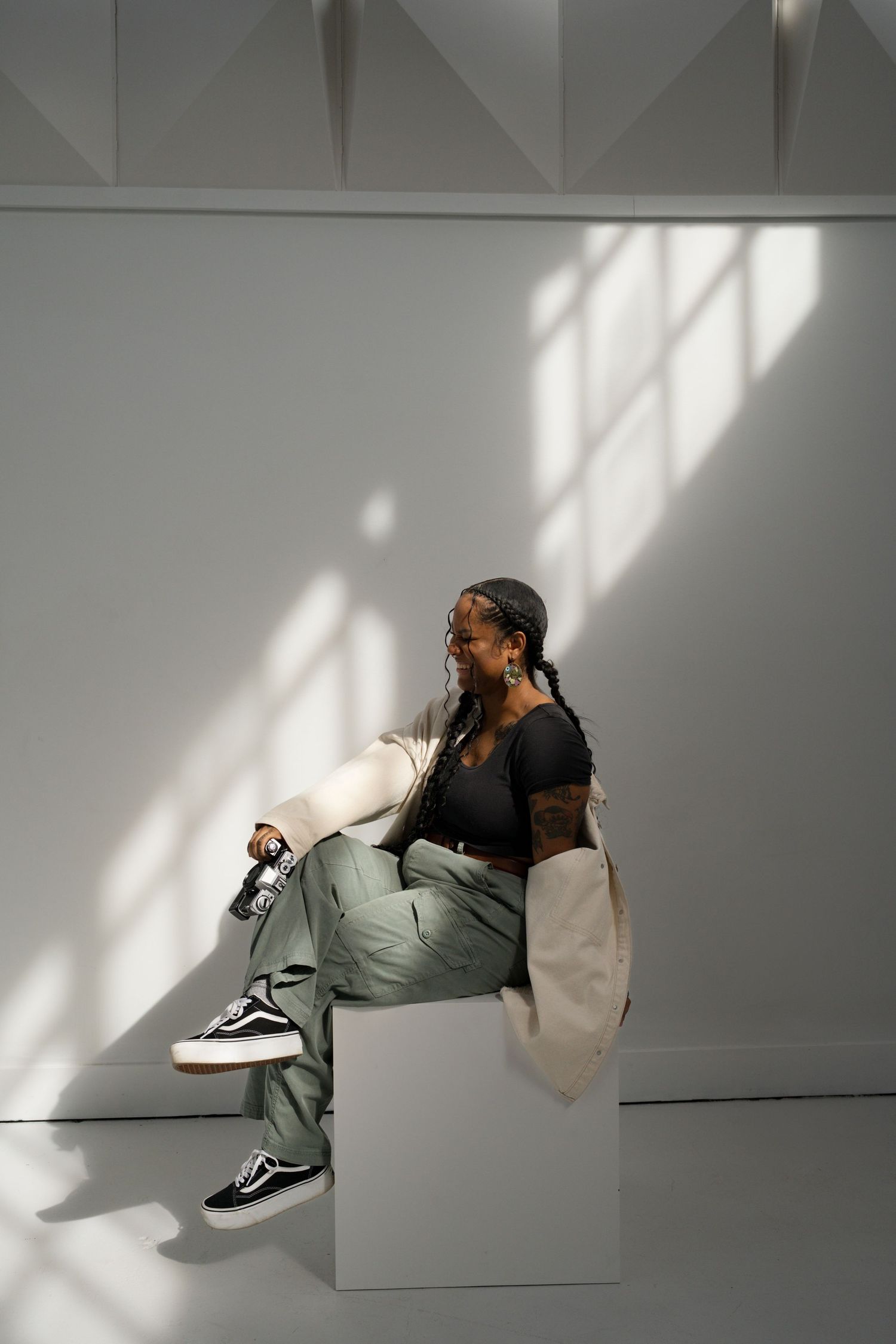 A Black woman with braids sits on a white cube holding a film camera, wearing a white jacket and sage green cargo pants.