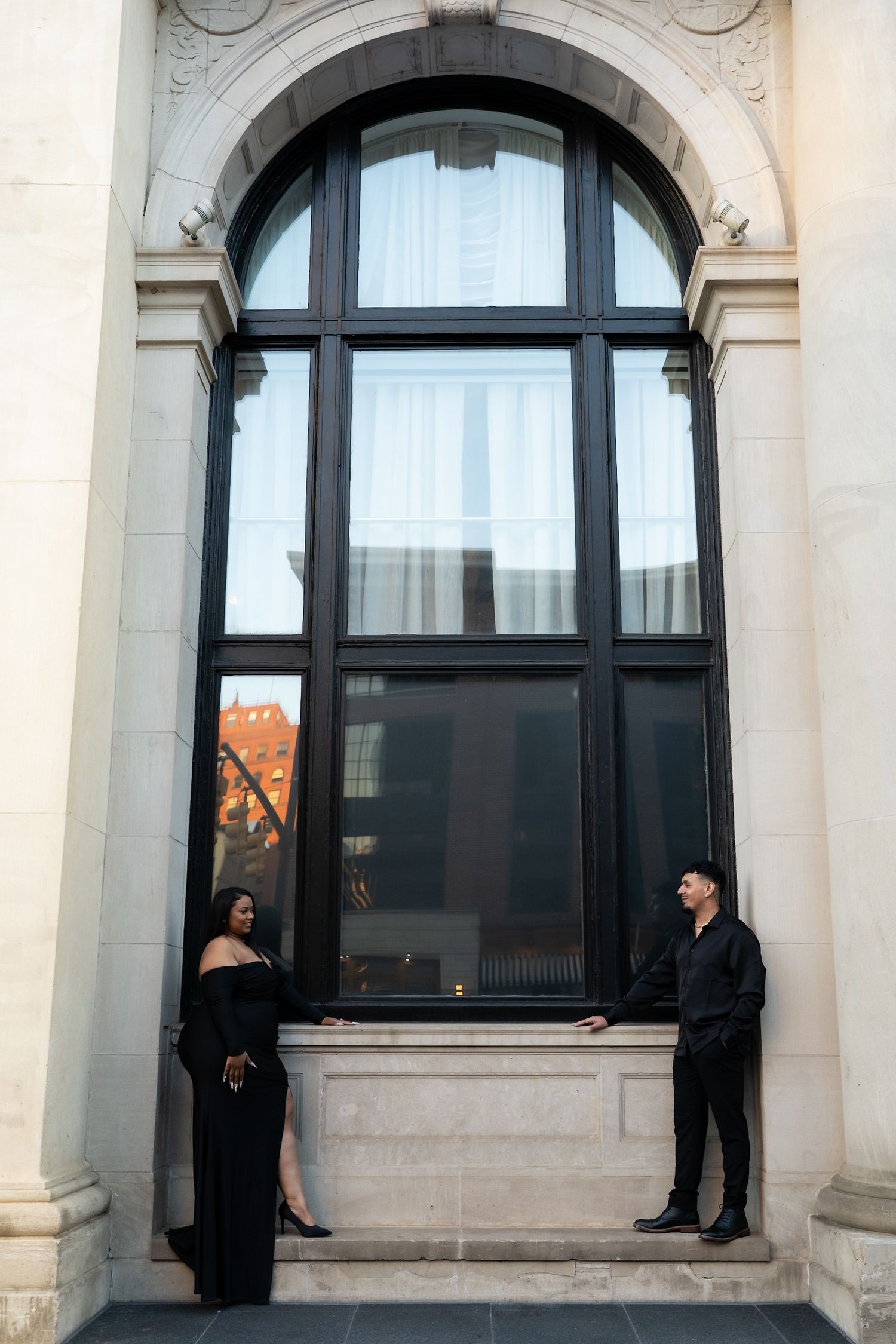 Stylish couple in black outfits standing apart before a large arched window on a classical building exterior.
