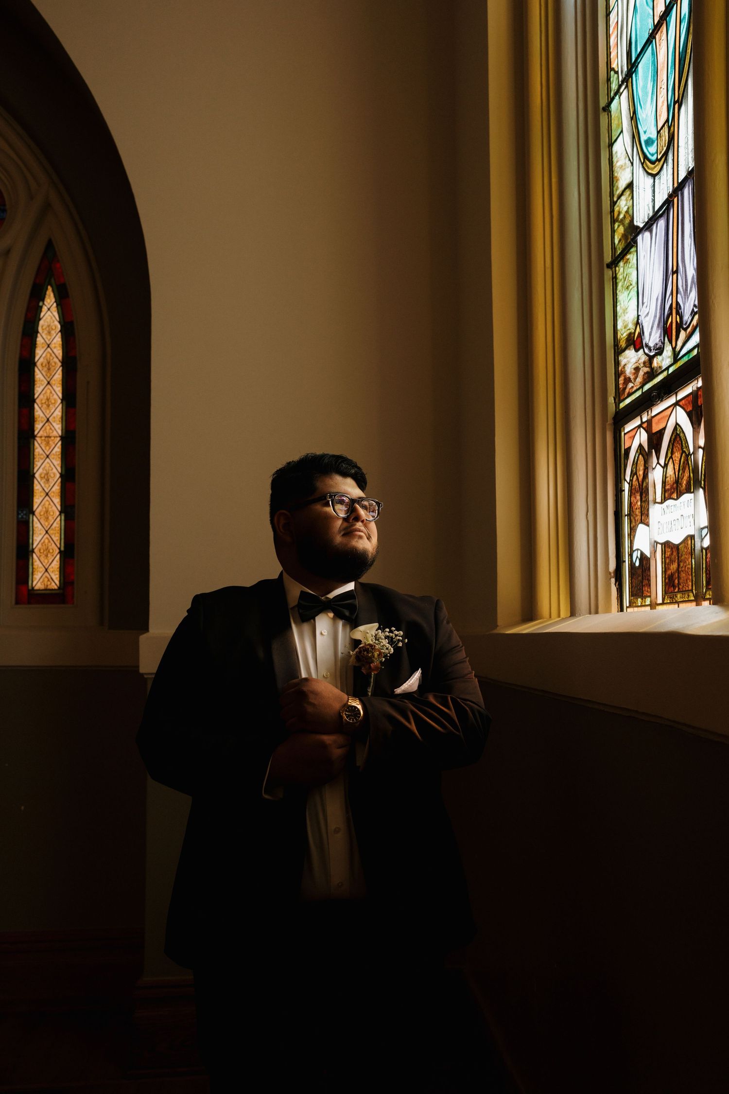 Groom in black tuxedo gazing upward beside a vibrant stained glass window in a church.
