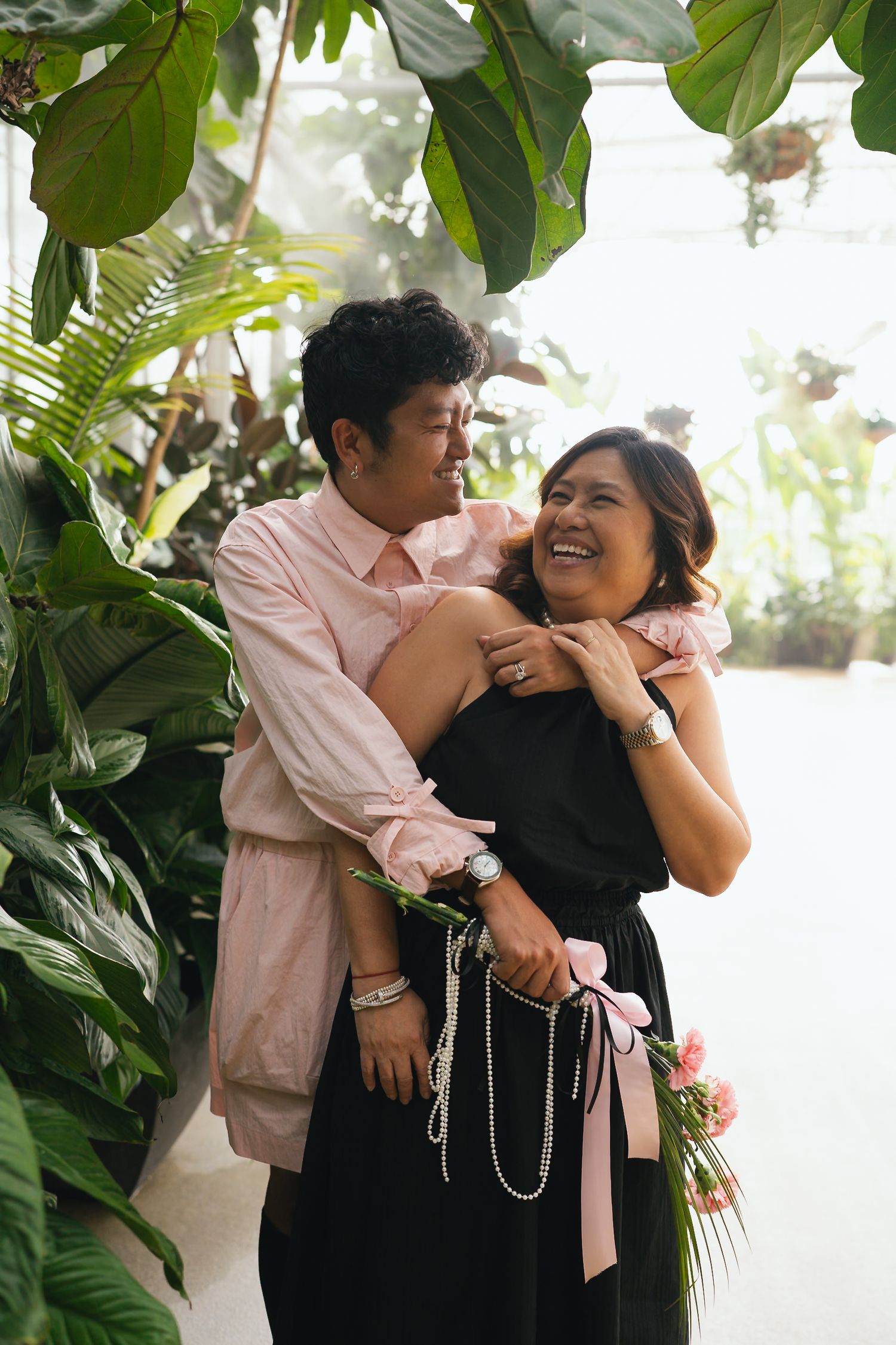 Two women laughing joyfully among tropical plants, one in pink shirt embracing other in black dress with pink bow.