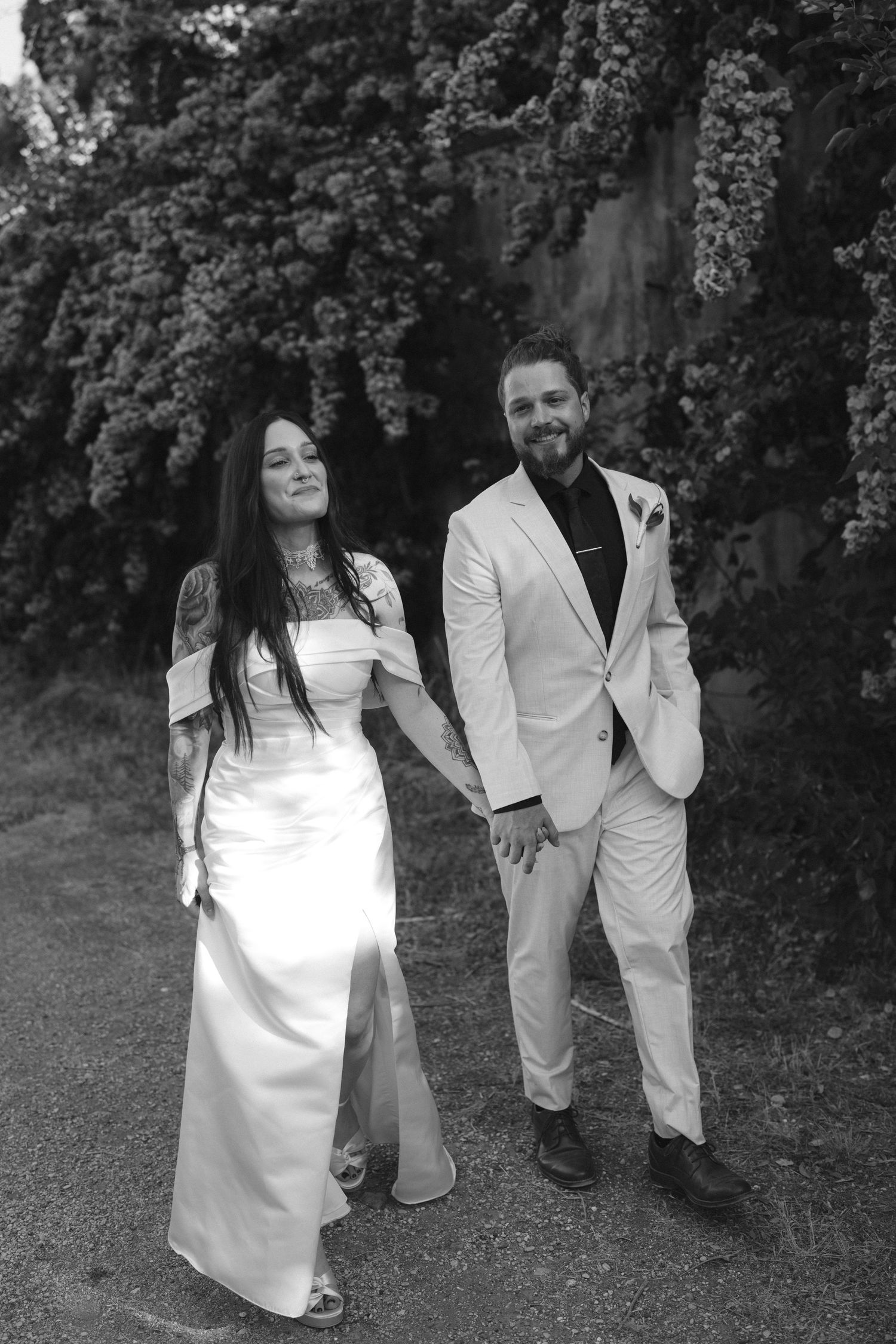Black and white photo of a bride in white gown and groom in white suit holding hands outdoors near lush greenery.