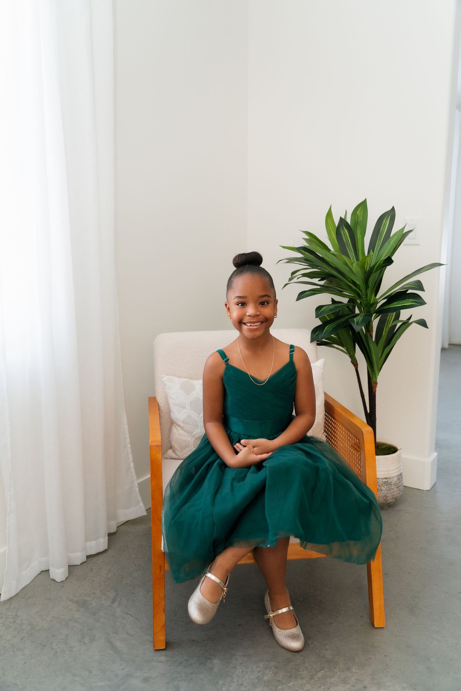 Young girl in emerald green tulle dress sits smiling in a wooden chair beside an indoor plant.