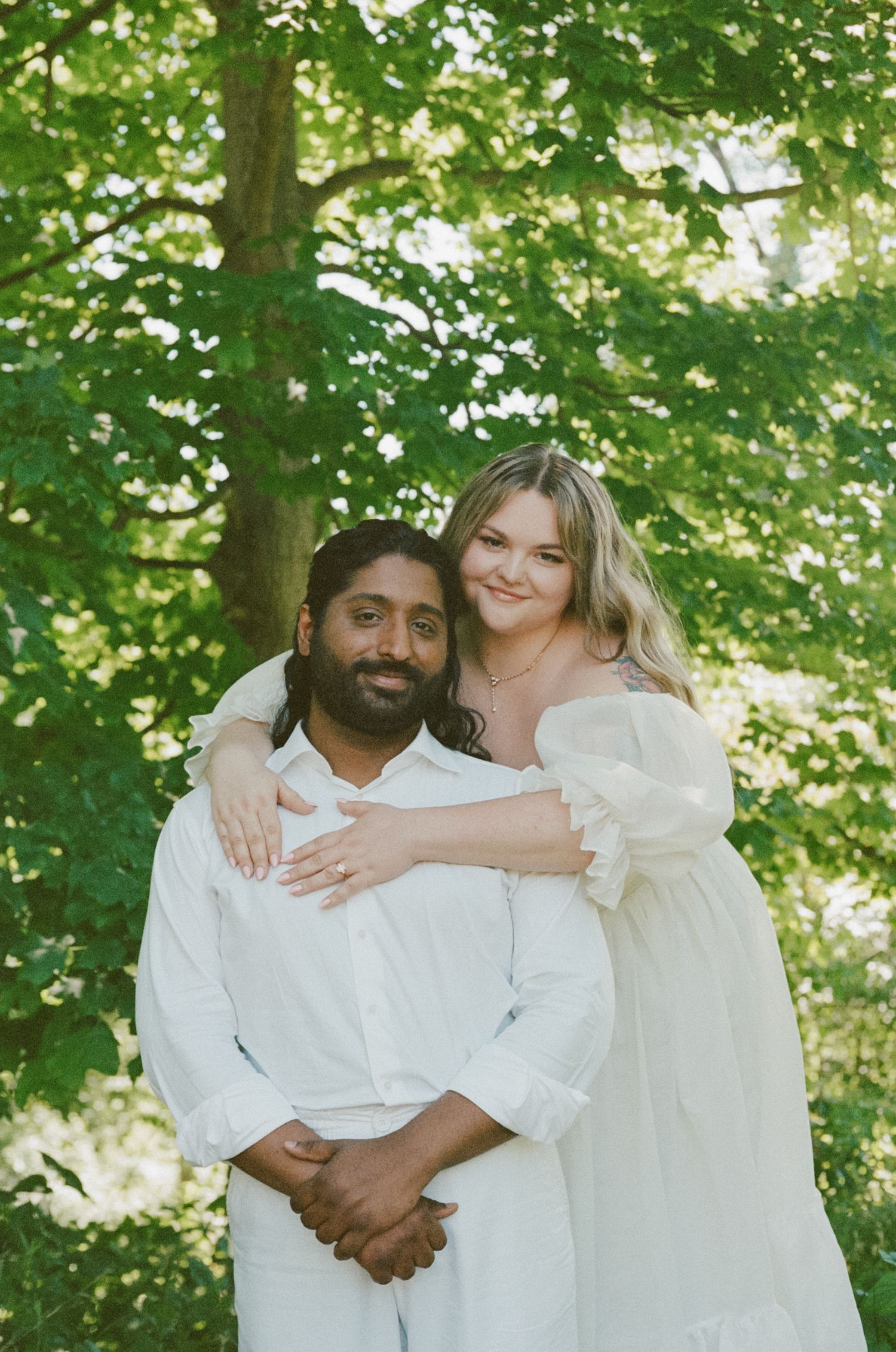 Couple embracing outdoors in white outfits surrounded by lush green trees.