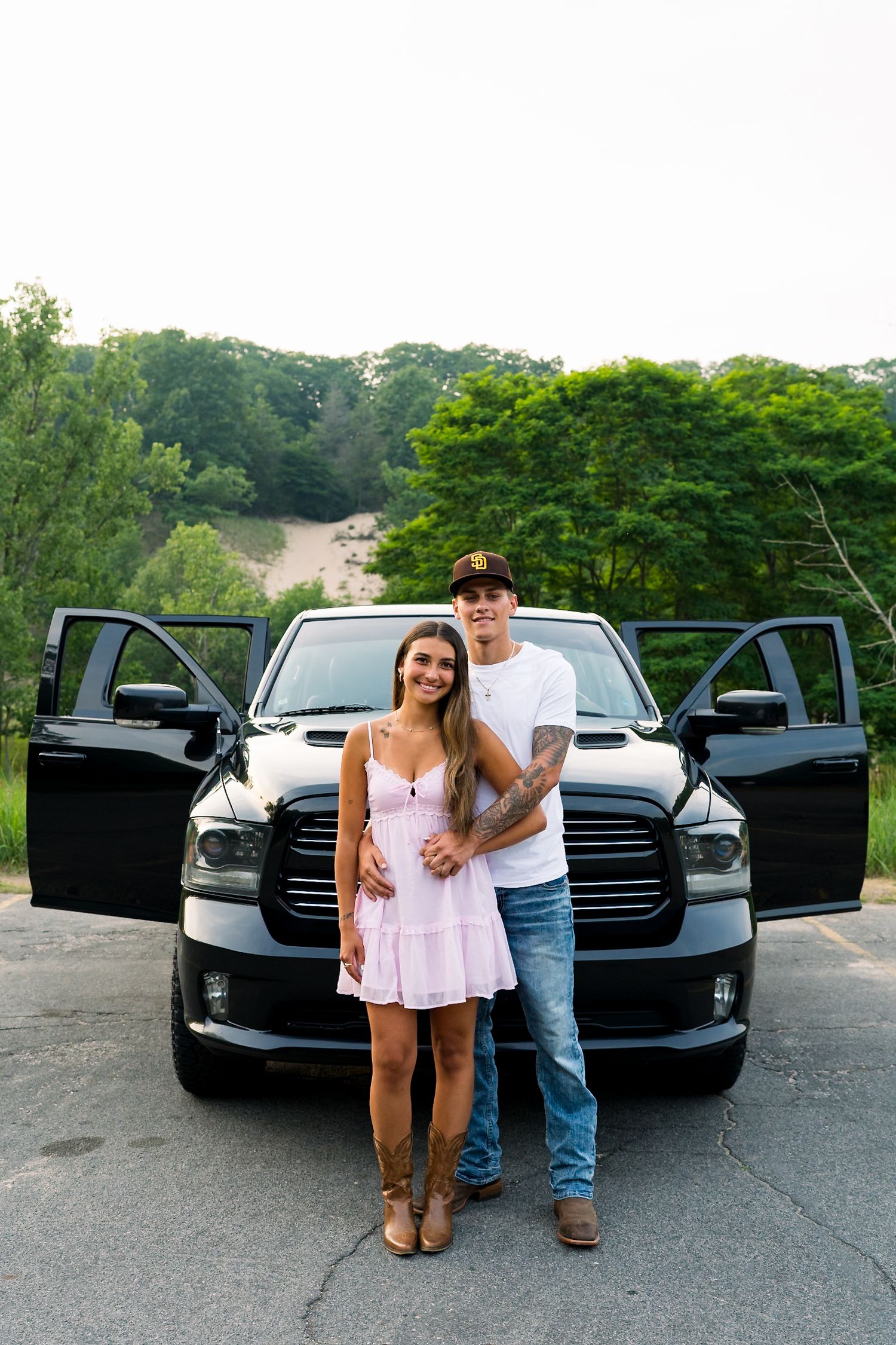 Young couple in pink dress and jeans posing in front of black Dodge Ram pickup truck with open doors outdoors.