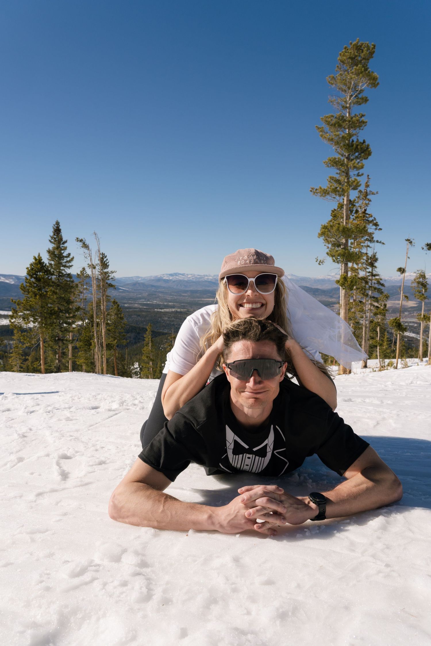 Couple posing in snow on a sunny mountain slope surrounded by pine trees and clear blue sky.